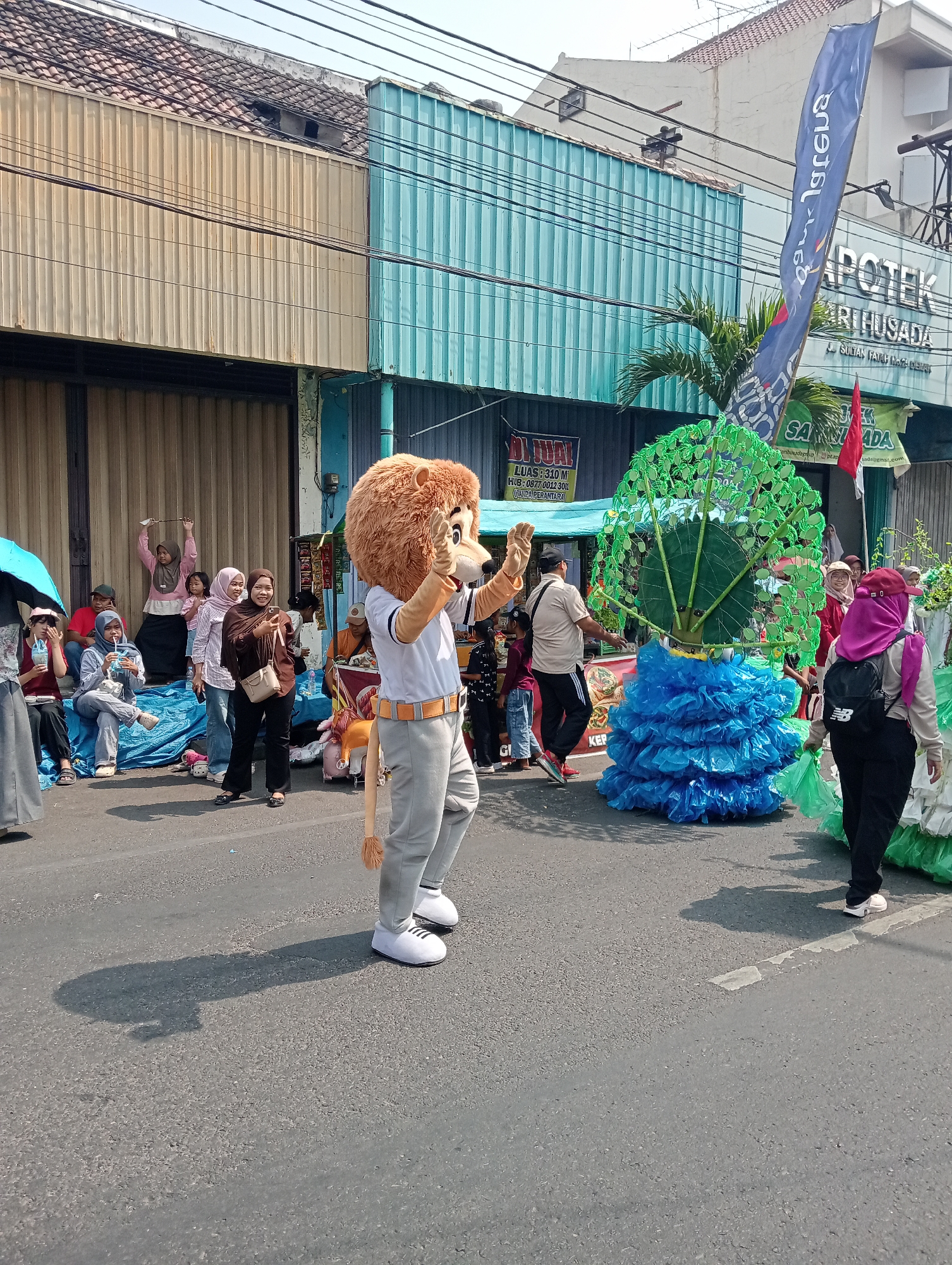 The image depicts a street scene in Bintoro, Indonesia, possibly during a parade or festival. A person in a lion mascot costume is walking down the street, waving. To the right of the mascot is a large, elaborate float resembling a tree with green leaves and a blue, ruffled base. Spectators are gathered on the side of the street, some sitting on blue tarps and others standing. One woman is holding up a phone to take a picture. In the background, there are buildings with corrugated metal siding, a blue building with a turquoise stripe, and a pharmacy sign that reads "APOTEK". A blue flag with white lettering is also visible, along with a red flag. The street is paved and appears to be sunny, with shadows cast by the people and floats.