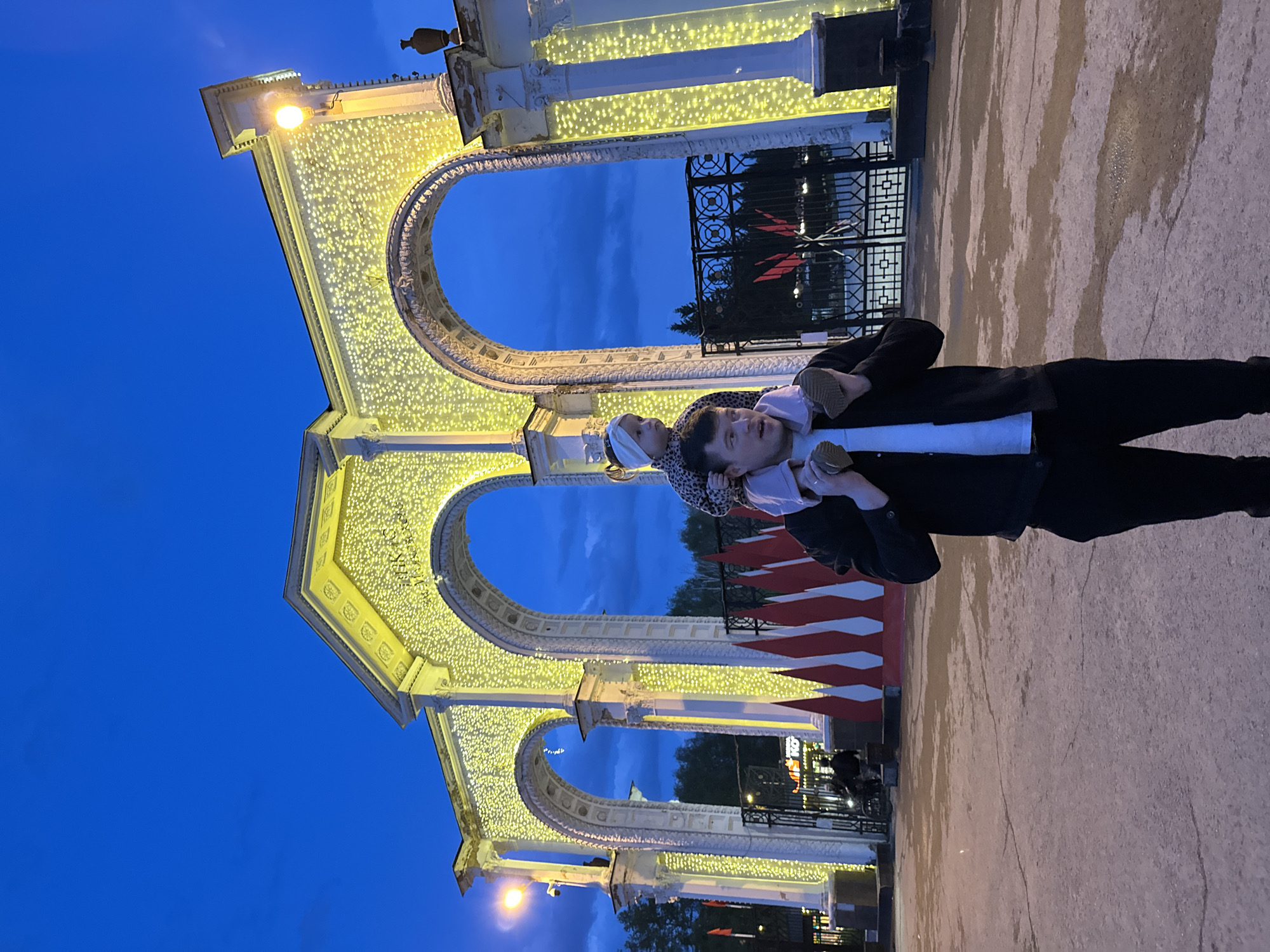 A father carries his young child on his shoulders in the city of Yekaterinburg, Russia, in front of an illuminated triple arch structure. The arches are decorated with strings of lights. The father is wearing a black suit jacket and pants, while the child is wearing a patterned jacket. Behind the father and child is a metal gate with a red symbol on it, as well as red and white flags. The sky is a deep blue, suggesting it is dusk or night. The ground appears to be wet, reflecting the lights of the arches.