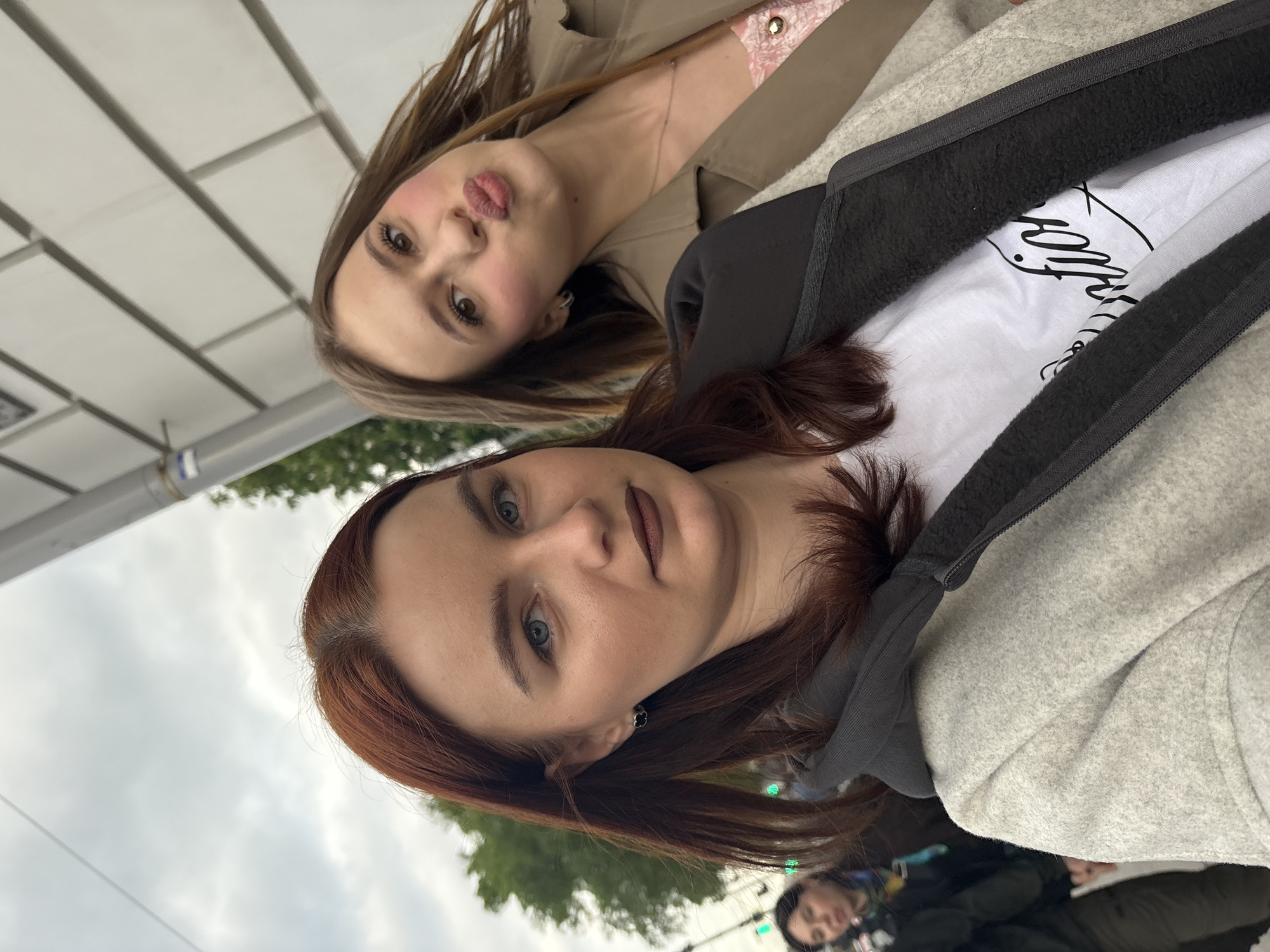 Two women pose for a selfie. The woman in the foreground has auburn hair, blue eyes, and is wearing a grey sweatshirt over a white t-shirt with black writing. The woman in the background has light brown hair and is wearing a tan trench coat over a floral print top. They are standing near a bus stop or shelter. Street lights and foliage are visible in the background, suggesting an urban environment. The overall setting appears to be Saint Petersburg, Russia, based on the architectural style of the bus stop and typical Russian streetscape elements.