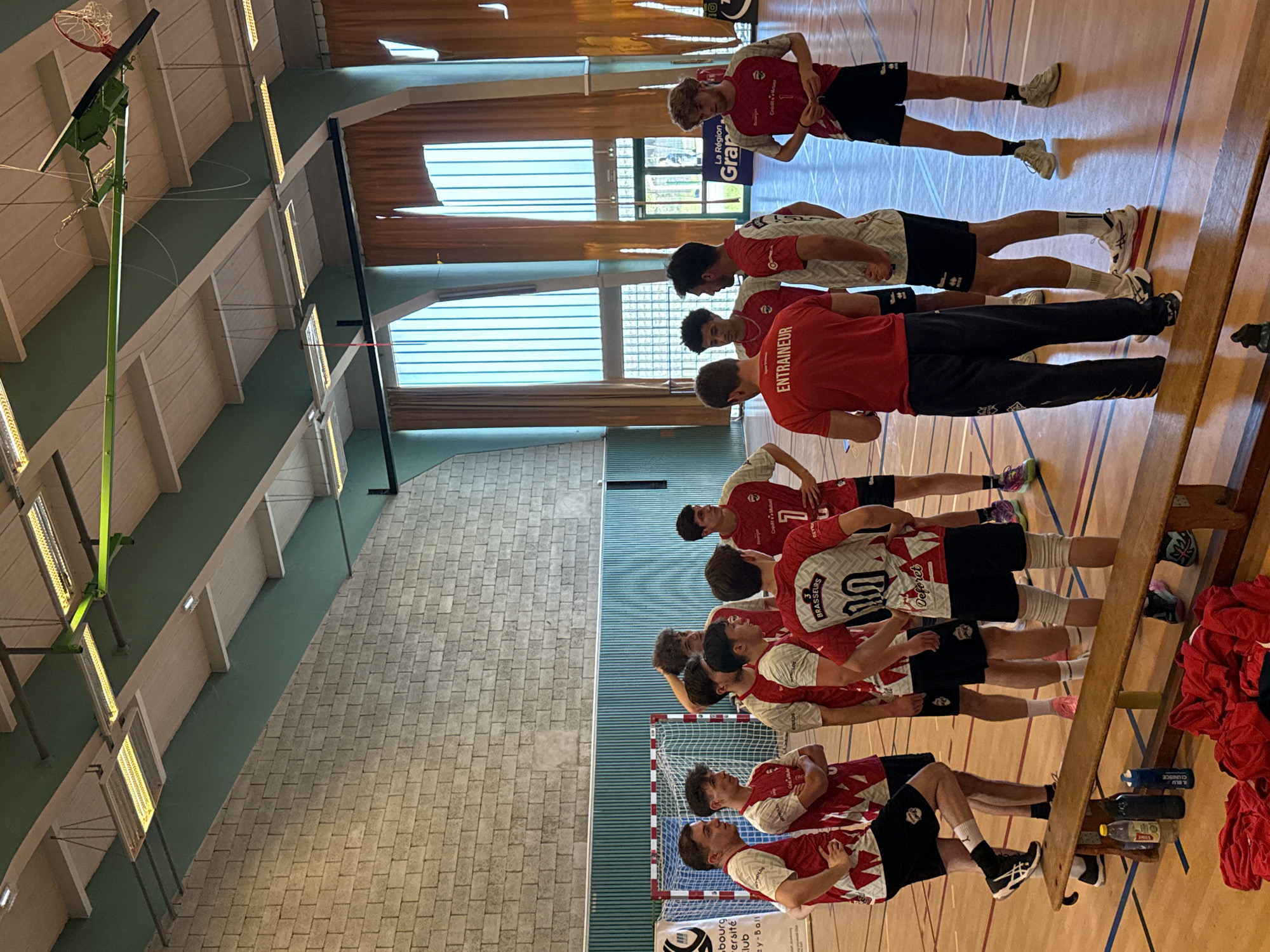 A photo taken inside a gymnasium depicts a handball team, dressed in white and red jerseys and black shorts, facing away from the camera, lined up in a gymnasium. The person in the middle of the line is wearing a red shirt that says "ENTRAINEUR" (coach) in white letters. The scene is likely at Strasbourg University Club, a handball club from Strasbourg, France.