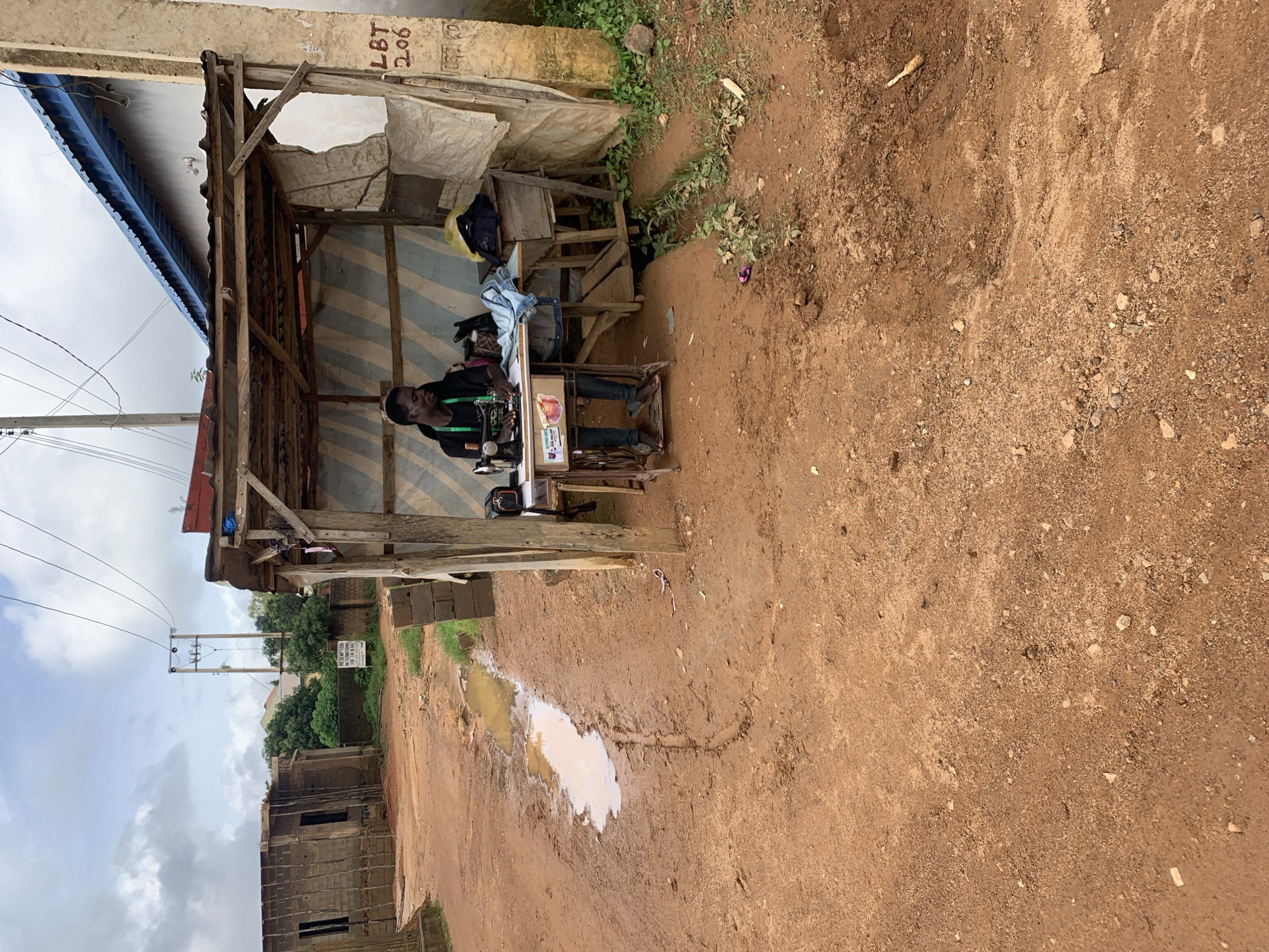 In Jos, Nigeria, a man is seen working as a tailor in a makeshift outdoor stall. He is seated at a vintage sewing machine, actively engaged in his work. The stall is constructed from wood and corrugated iron, with a fabric covering providing shelter. To the left of the stall, a muddy puddle reflects the overcast sky, indicating recent rainfall. The ground is predominantly red earth, typical of many regions in Nigeria. In the background, unfinished buildings and utility poles suggest a developing urban landscape. The overall scene depicts a common sight of small businesses operating in informal settings within the city.