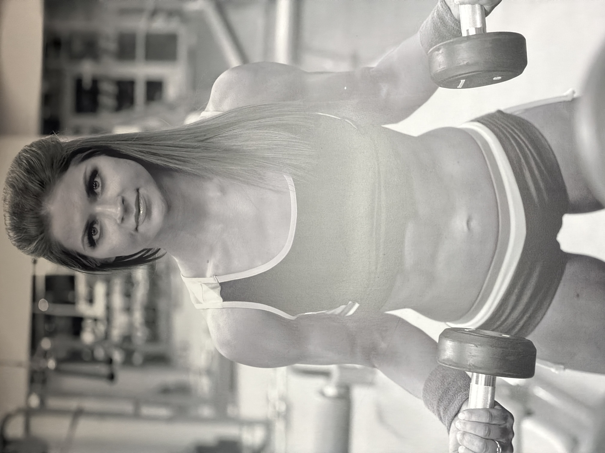 A black and white photograph depicts a woman posing in what appears to be a gym setting. The image is oriented with the woman lying horizontally, but should be viewed as if she is standing or sitting upright, looking towards the viewer.
The primary subject is a young to middle-aged woman with long, dark hair that falls over her shoulders. She has a direct gaze towards the camera, with a hint of a smile or smirk, conveying confidence. She appears to have an athletic build.
She is wearing athletic apparel, including a sports bra or crop top and shorts featuring prominent stripes. A wristband is visible on her right wrist. In each hand, she holds a dumbbell, resting them slightly below her waist.
The background is softly blurred, showing indistinct shapes and forms consistent with gym equipment or the interior architecture of a fitness facility. The lighting is soft and even, highlighting the subject while obscuring specific details of the environment. There is no visible text in the image. The overall impression is that of a posed shot, possibly for a fitness advertisement or personal portfolio, rather than a candid moment during a workout.