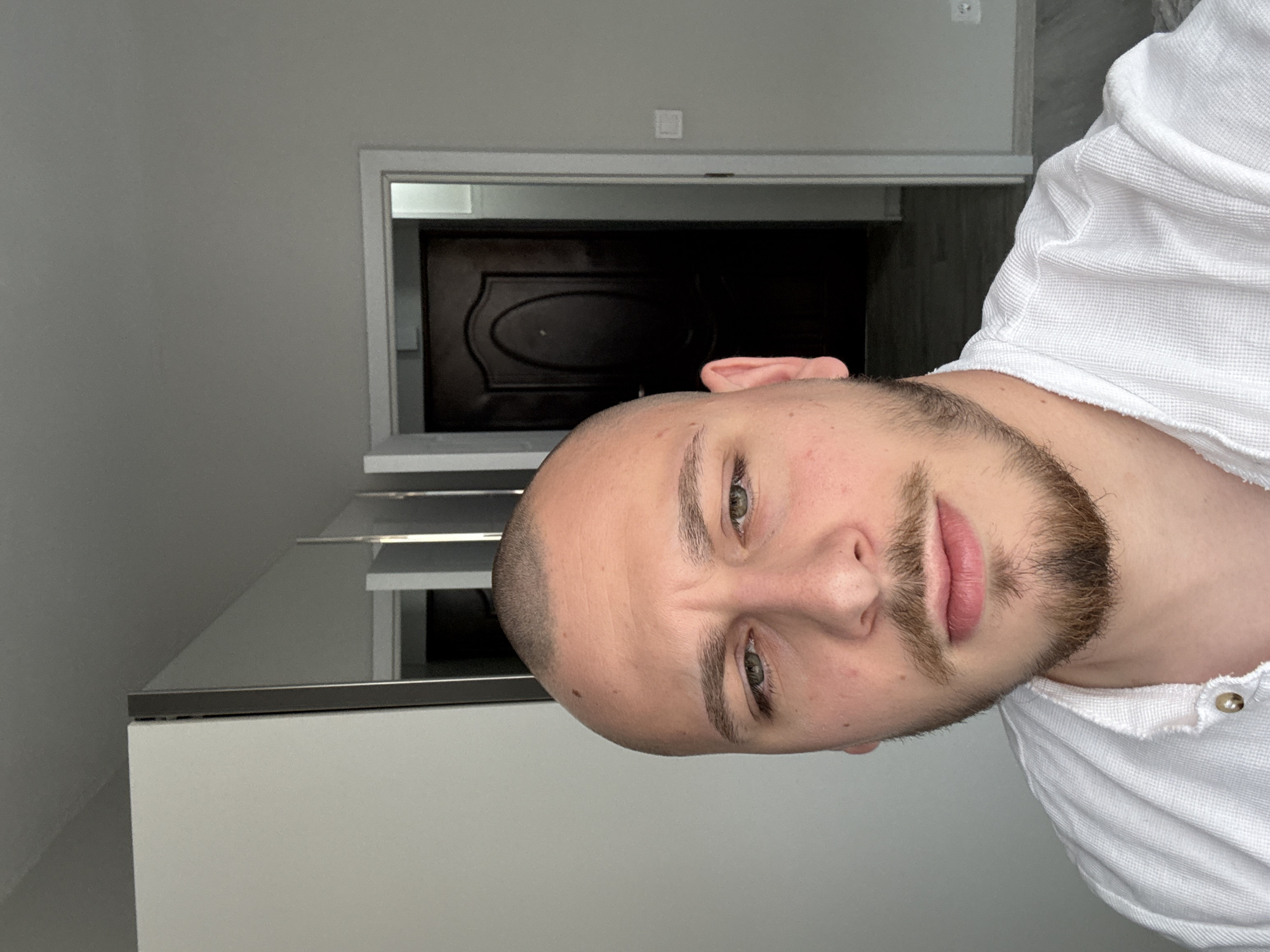 The scene depicts an adult male subject in a residential interior located in Stara Zagora, Bulgaria. The male, with a shaved head, green eyes, and a short brown beard and mustache, faces the camera directly with a neutral expression. He is wearing a white, textured, collared garment, possibly a waffle-knit shirt or robe, with two gold-colored buttons visible. The background features light gray walls. To the subject's left, a white wardrobe with reflective mirrored doors is visible, partially reflecting the room's interior. To the subject's right and extending behind him, a dark brown, paneled wooden door is partially open within a white frame, revealing a dimly lit hallway or adjacent room with a dark floor. A white electrical outlet is affixed to the lower right wall.
