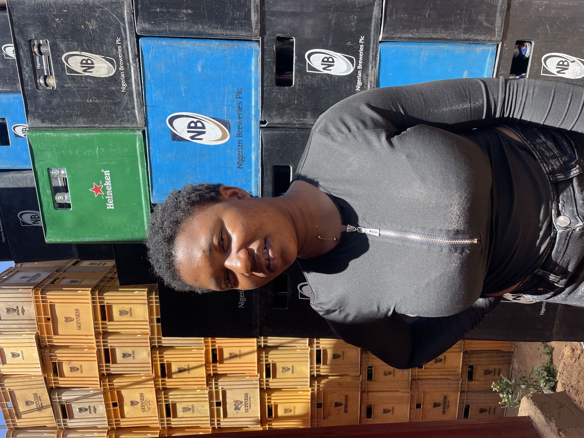 The image shows a person standing in front of a stacked wall of beer crates in Gura Top, Nigeria. The person, appearing to be female, is wearing a black, zipper-front shirt and dark jeans. The crates behind them are various colors and brands, including Guinness (tan), Heineken (green), and Nigerian Breweries (black and blue). Some crates appear to be full of bottles, while others are empty. The ground appears to be dirt and a small plant is visible near the bottom right corner of the image.