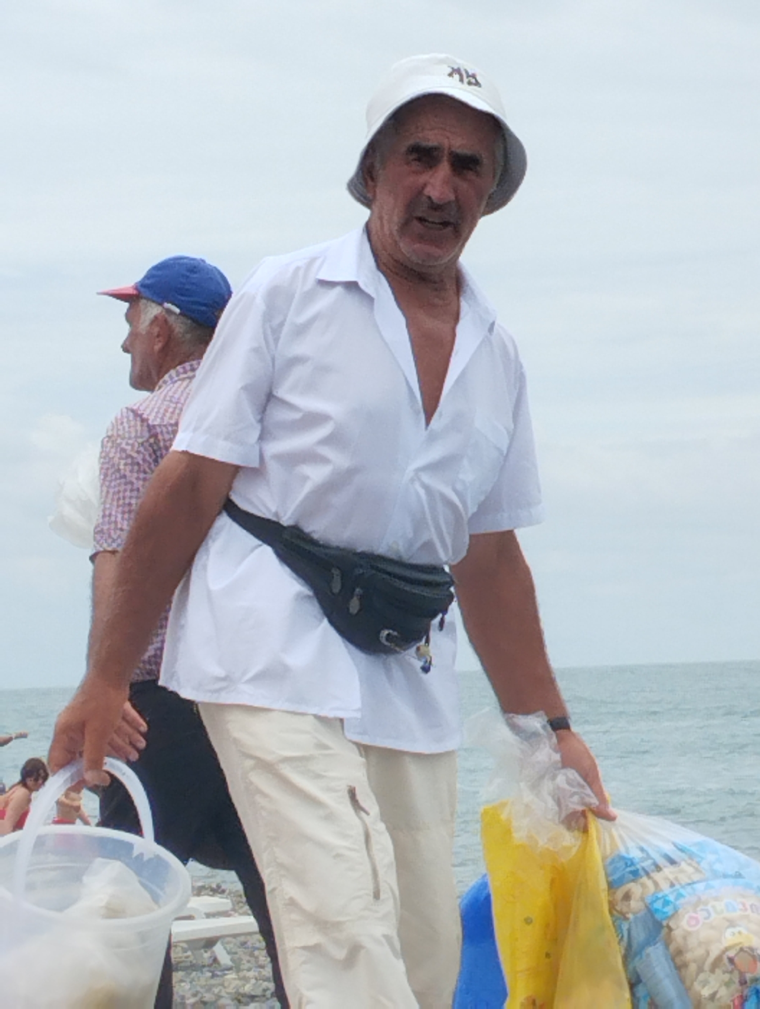 A medium shot captures an older man in a white hat and white short-sleeved open-collar shirt, standing on a beach. He wears a black fanny pack around his waist and light-colored pants. In his right hand, he holds a clear plastic bucket, and in his left, a plastic bag filled with yellow and blue items. Behind him, another man in a blue cap and checkered shirt is visible, and further in the background, two women in swimwear are on the beach. The scene is set against a backdrop of the ocean and an overcast sky.