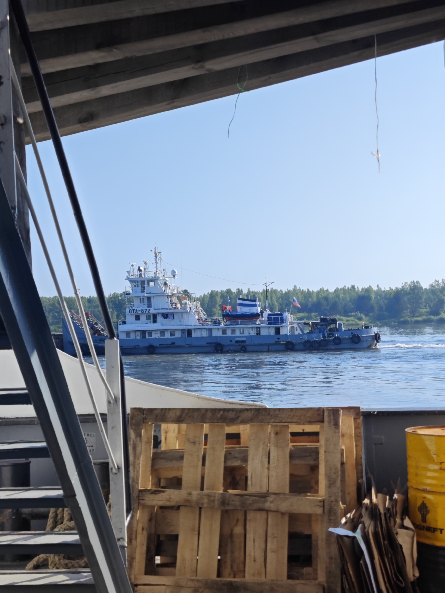 This is a medium shot, taken from a low angle, of a canal or river in Samkovo, Russia. The foreground is dominated by the deck of a vessel, with wooden pallets and a yellow barrel visible on the right. A portion of a ship's stairs is also on the left. In the mid-ground, a large white and blue ship with the identification "OTA-872" is moving down the waterway. The ship has several decks and a Russian flag flying. The background consists of a dense green forest lining the opposite bank of the river, under a clear blue sky. The lighting suggests it is daytime, with sunlight illuminating the scene. There are no people visible in the image.