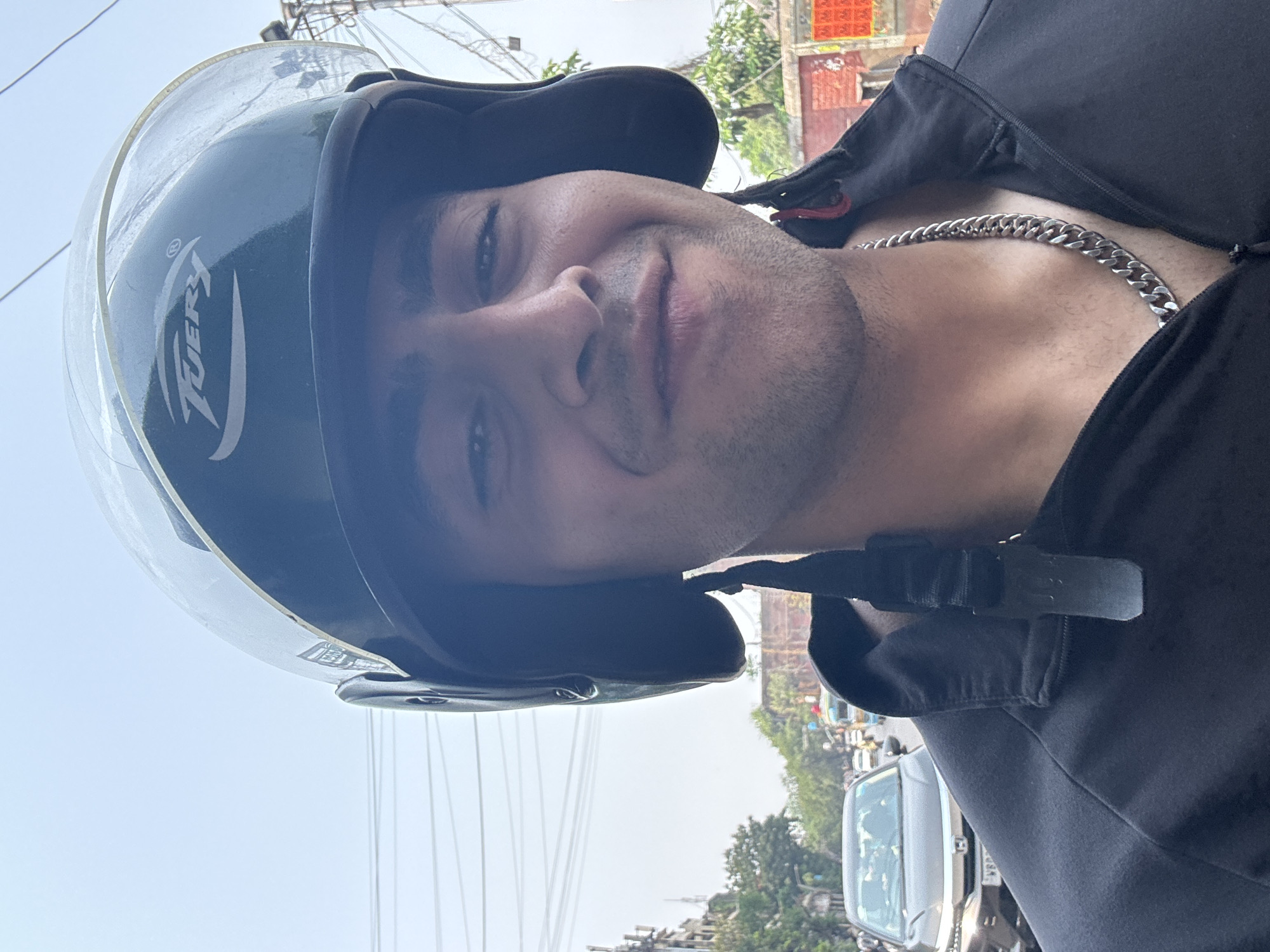 A man wearing a helmet and a black jacket poses for a selfie. He is also wearing a silver chain. Outside the frame, there is a street with trees and vehicles. This photograph was taken in Kolkata, India.