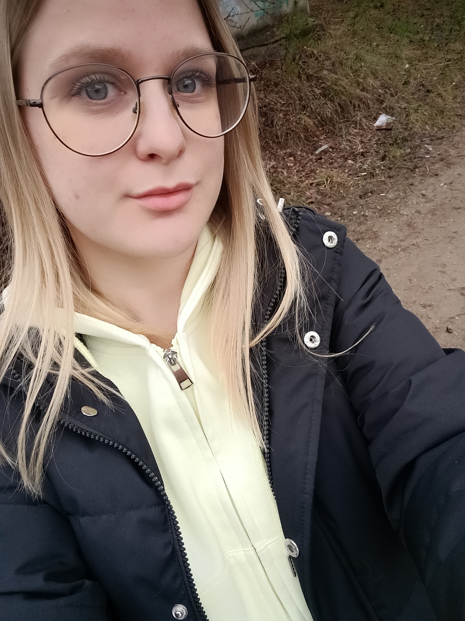 This is a close-up, chest-up selfie of a young woman taken outdoors in Noviy, Russia. She has long, straight blonde hair, which partially covers her shoulders and jacket, and light blue eyes. She wears large, round, dark metal-framed glasses and has a neutral expression while looking directly at the camera.
She is dressed in warm attire, including a dark navy or black puffer jacket worn open over a light yellow hooded sweatshirt. The sweatshirt has a prominent silver-toned zipper pull. Silver snap buttons are visible on the front placket of her jacket.
The background consists of an undeveloped outdoor area. To the right, there is a dirt or gravel path, while the left features an uneven patch of dry, brown grass, leaves, and some sparse vegetation. Small pieces of white litter are visible scattered on the ground in the mid-background. The soft, even lighting suggests an overcast day or late afternoon, and the warm clothing indicates cool weather. No text is visible in the image.