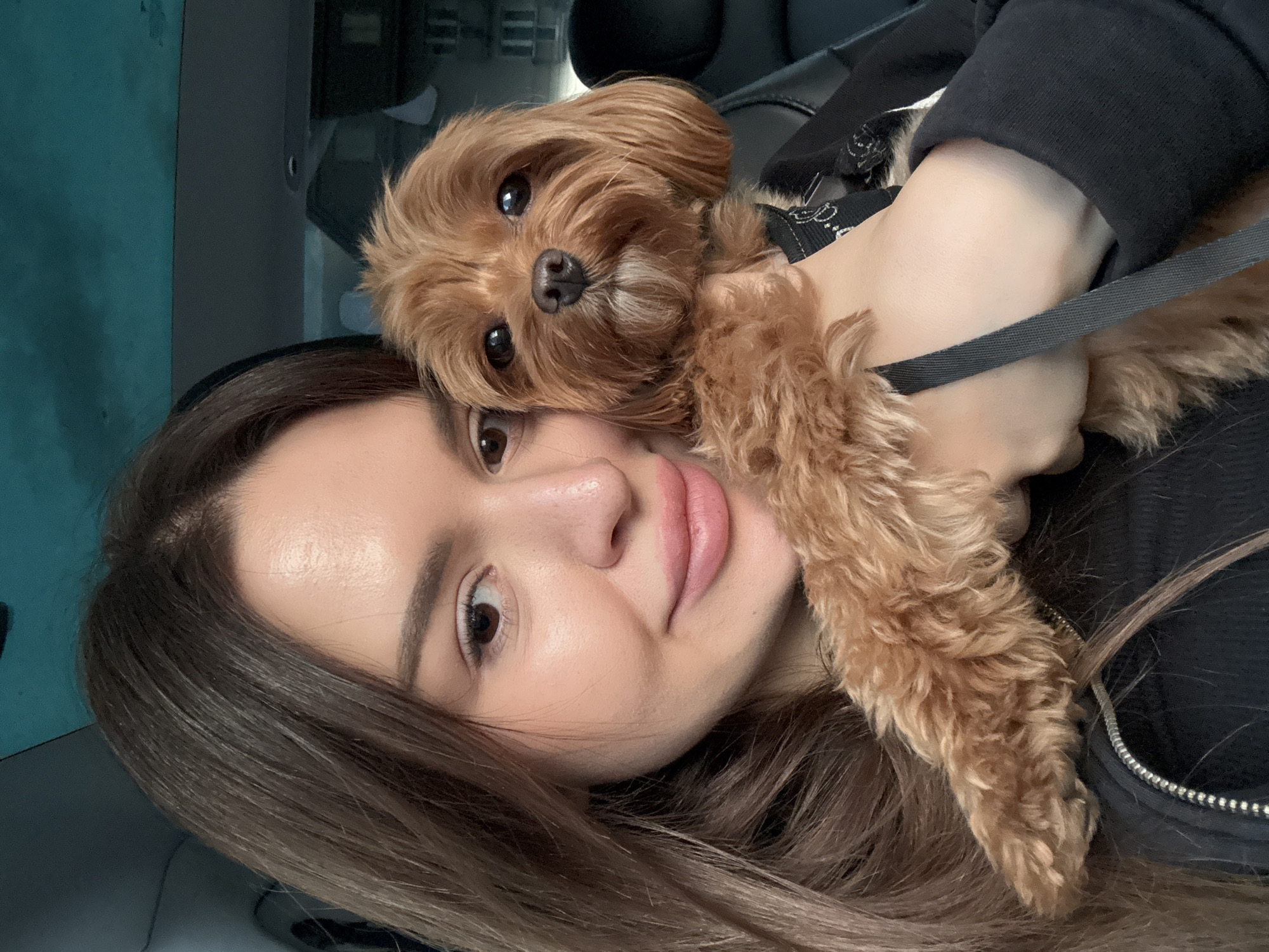 A young woman with long, dark brown hair and light makeup is holding a small, light brown dog with fluffy fur. She is in a car, as indicated by the headrests and ceiling visible in the background. The woman is looking at the camera with a slight smile, and the dog is looking directly at the camera with its mouth slightly open. The dog is wearing a black collar. The lighting suggests it is daytime. The image does not contain any identifiable landmarks or clues that would indicate a specific location within Prague, Czechia.