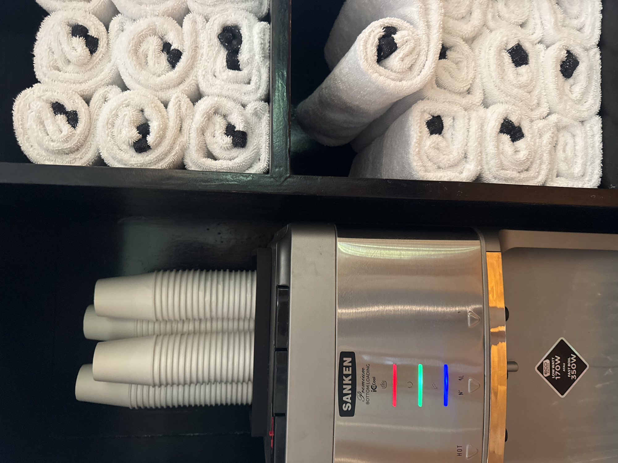 This image shows a shelf with rolled white towels in the top compartments. The towels in the left compartment are rolled neatly, while those in the right compartment are more loosely stacked. Below the shelf is a "Sanken" brand water dispenser with paper cups stacked to its left. The dispenser has LED indicators with the words "HOT", "NAL" at the bottom, and "ECO" next to the "LOW WATT 170W" label. The setting is likely in Kuta Selatan, Indonesia, based on the brand of the water dispenser.