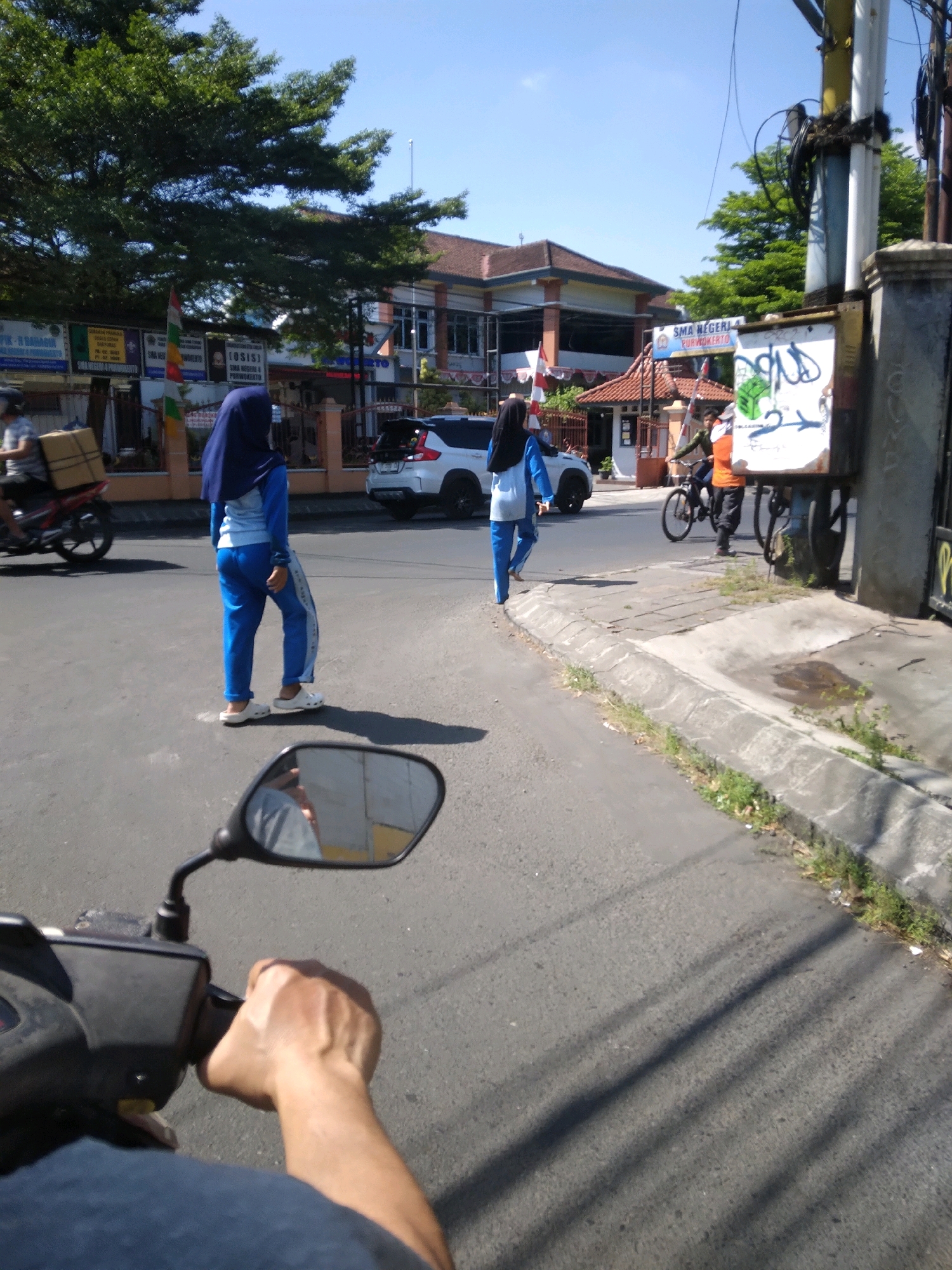 In Purwokerto, Indonesia, a street scene unfolds under a bright blue sky. The foreground shows a person's hand gripping the handlebar of a motorcycle, with a partial view of the motorcycle's dashboard and a rearview mirror reflecting a person wearing a grey shirt. On the street, two female students wearing blue uniforms and headscarves walk in the same direction. The student closer to the viewer is on the left side of the frame, facing away from the camera. The other student is further down the road on the right side, also facing away. Behind them, a white SUV is parked on the right side of the street. A man in an orange shirt and dark pants is standing next to a bicycle near a utility pole on the sidewalk. The background features a building with a red-tiled roof, possibly a school, identified by signage reading "SMA NEGERI 4 PURWOKERTO." Several trees with lush green foliage line the street. The environment appears to be a typical urban street with sidewalks, a paved road, and a clear sky.