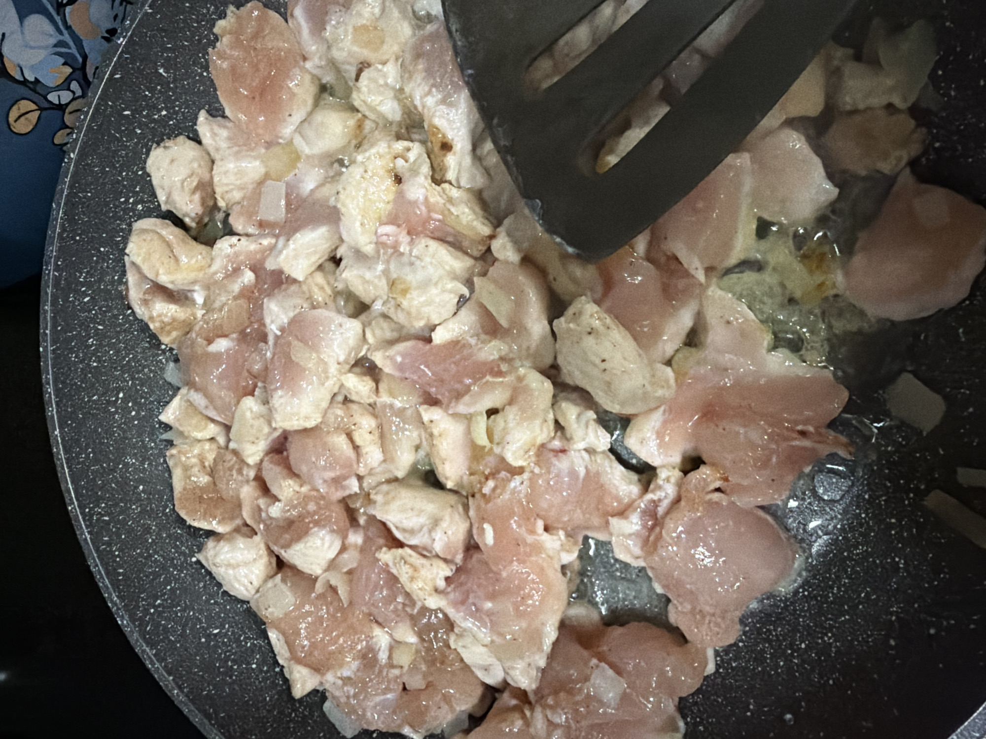 A close-up overhead shot shows cubed chicken pieces in a black non-stick frying pan. The chicken is raw and has a light pink hue, with some pieces beginning to turn opaque. There are also small pieces of white onion interspersed with the chicken. The pan is speckled with white dots. A dark gray spatula is visible on the right side of the frame, with its edge resting on some of the chicken pieces. The background is blurred and dark. There are no people or animals visible in the image. The scene appears to be a kitchen setting, with the focus on the raw ingredients being prepared for cooking. There is no information in the image that would suggest a location in Budva, Montenegro.