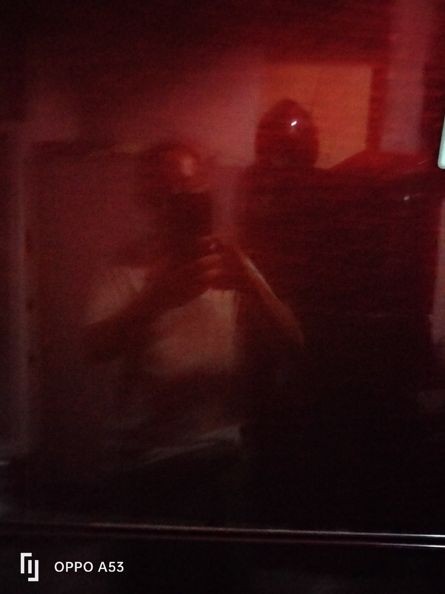 The image, taken in Denpasar, Indonesia, is a dark, blurry reflection on a red, glossy surface, likely a refrigerator door. The reflection shows two figures. On the left, a person is holding a smartphone and appearing to take a selfie. The person's face is indistinct. To the right, another figure, possibly wearing a helmet or hat with a light-colored marking, is visible. Behind the figures, a rectangular light source is reflected. The bottom of the image features a white watermark with the text "OPPO A53". The overall impression is of a dimly lit environment with reflections creating a somewhat abstract composition.