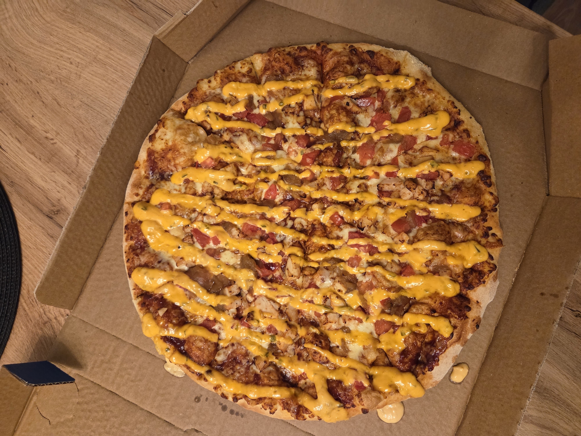 A pizza is inside a cardboard pizza box. The pizza is round with a golden crust. It is topped with cheese, chopped tomatoes, and chicken pieces, drizzled with a yellow sauce. The pizza box is open and is sitting on a wooden surface. There are no indications of location within Varna, Bulgaria, or any people present.