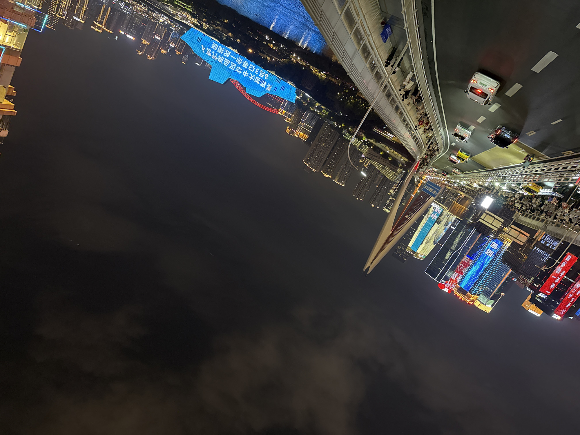The image depicts a nighttime city scene in Yuzhong District, China, characterized by illuminated buildings and a river. A bridge, likely a pedestrian walkway, overlooks a multi-lane road with several vehicles, including cars and what appear to be taxis. Along the walkway, numerous people are visible, observing the city lights and the river below. The buildings in the background are brightly lit with a variety of colors and illuminated signage, suggesting commercial or residential structures. The river reflects the city's lights, adding to the vibrant ambiance. A prominent blue digital display on a building shows Chinese text, and a red arched structure, possibly part of another bridge or monument, is also visible. The sky is dark, with some clouds. The overall scene conveys a lively urban environment at night.