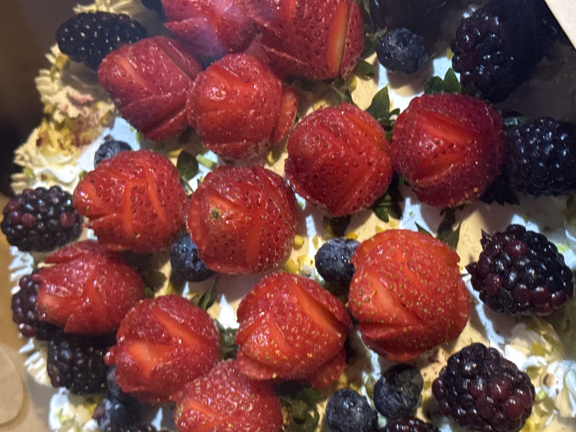 This close-up, overhead shot captures a vibrant assortment of fresh berries atop a dessert. The primary subjects are ripe strawberries, blackberries, and blueberries, all glistening with what appears to be water droplets, suggesting they have been recently washed or are naturally dewy. The berries are artfully arranged on a creamy white base, which is speckled with green pistachios, hinting at a cake or pastry.
The strawberries are whole and halved, displaying their characteristic red hue and seeded texture. The blackberries are plump and dark, with their individual drupelets clearly defined. The blueberries are smaller and round, adding a contrast in color and size. The overall scene is one of freshness and natural beauty, likely a dessert intended for enjoyment.
There are no people, text, or explicit location cues visible in the image. The lighting suggests an indoor setting, possibly a kitchen or a bakery, as there are no indications of natural daylight or outdoor elements. The focus is entirely on the textures and colors of the berries and the dessert. The image evokes a sense of sweetness and indulgence, a common association with such fruit-laden treats. The context of Chiang Mai City Municipality, Thailand, does not provide specific visual cues within the image itself, but it sets the geographical location where this dessert might be found.