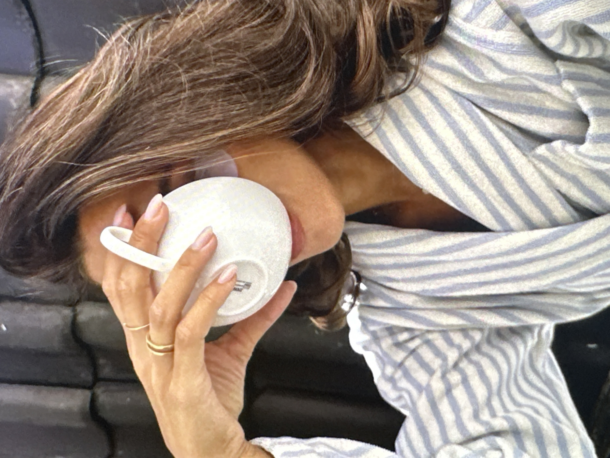A person with brown hair streaked with blonde is drinking from a white mug. The person's left hand is holding the mug and has two gold rings on the ring and middle fingers. The person is wearing a blue and white striped shirt or robe. The background appears dark. The location and city cannot be determined from this image.
