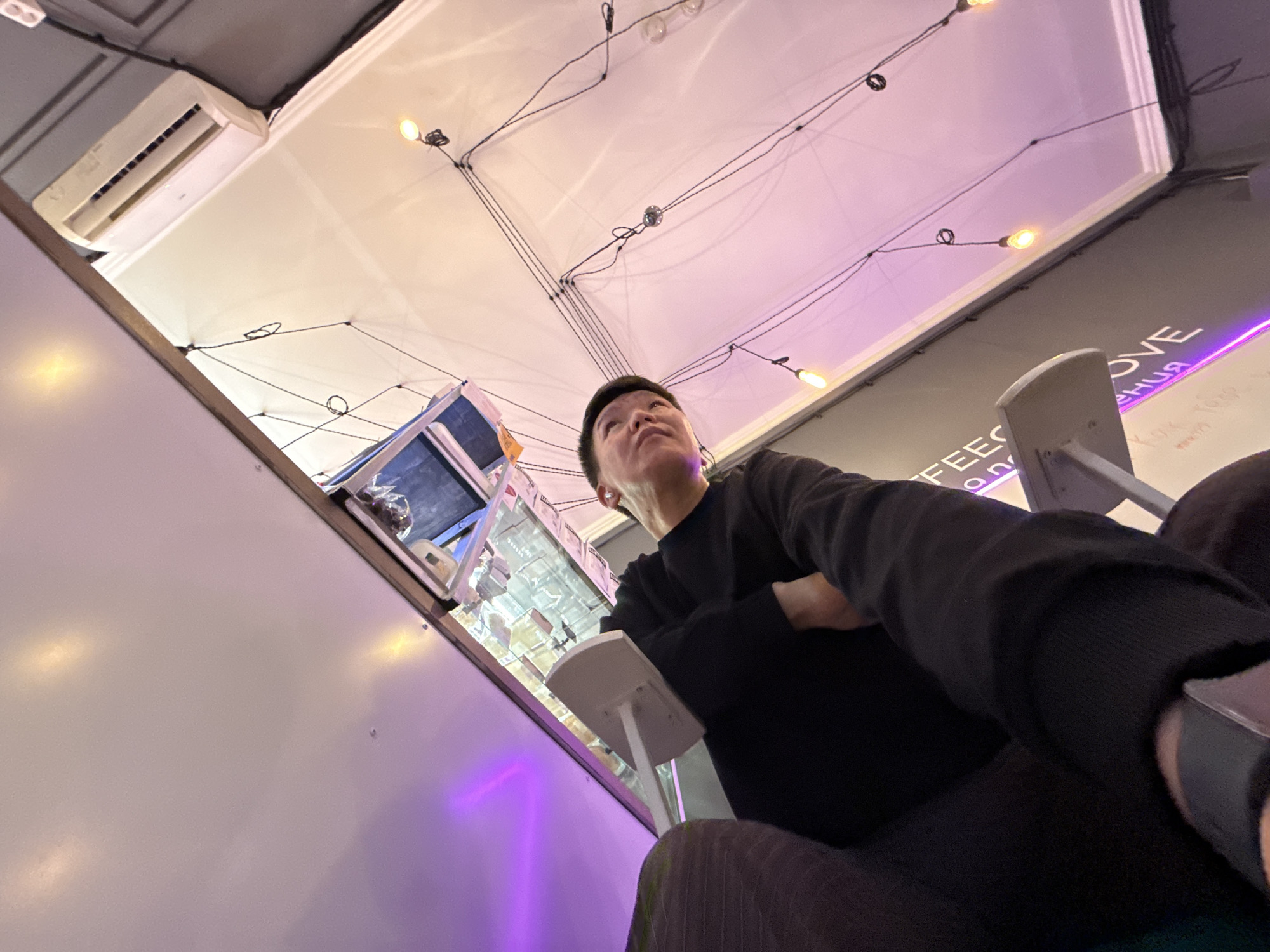 The image shows a low-angle shot of a young man sitting in what appears to be a cafe or restaurant. He is wearing a black long-sleeved shirt and black pants. He is looking upwards and to the right, with his mouth slightly open. A portion of the photographer's arm, wearing a black sleeve and a watch, is visible in the foreground.
The environment features a white ceiling with several strings of small, warm-toned Edison bulbs, as well as an air conditioning unit mounted high on the wall. There is a prominent purple light source to the left, casting a violet glow on the ceiling and a portion of the wall. A white table or counter is visible in the mid-ground, with a sign above it that appears to have text in both English and Russian, partially obscured. The English text includes "FEES" and "LOVE". The Russian text appears to be "КОМПЛЕКСНЫЕ" and "СЛУЖБА". The overall impression is a modern, casually lit interior space. It's not possible to determine the specific location within Moscow, Russia based on the visual information provided.