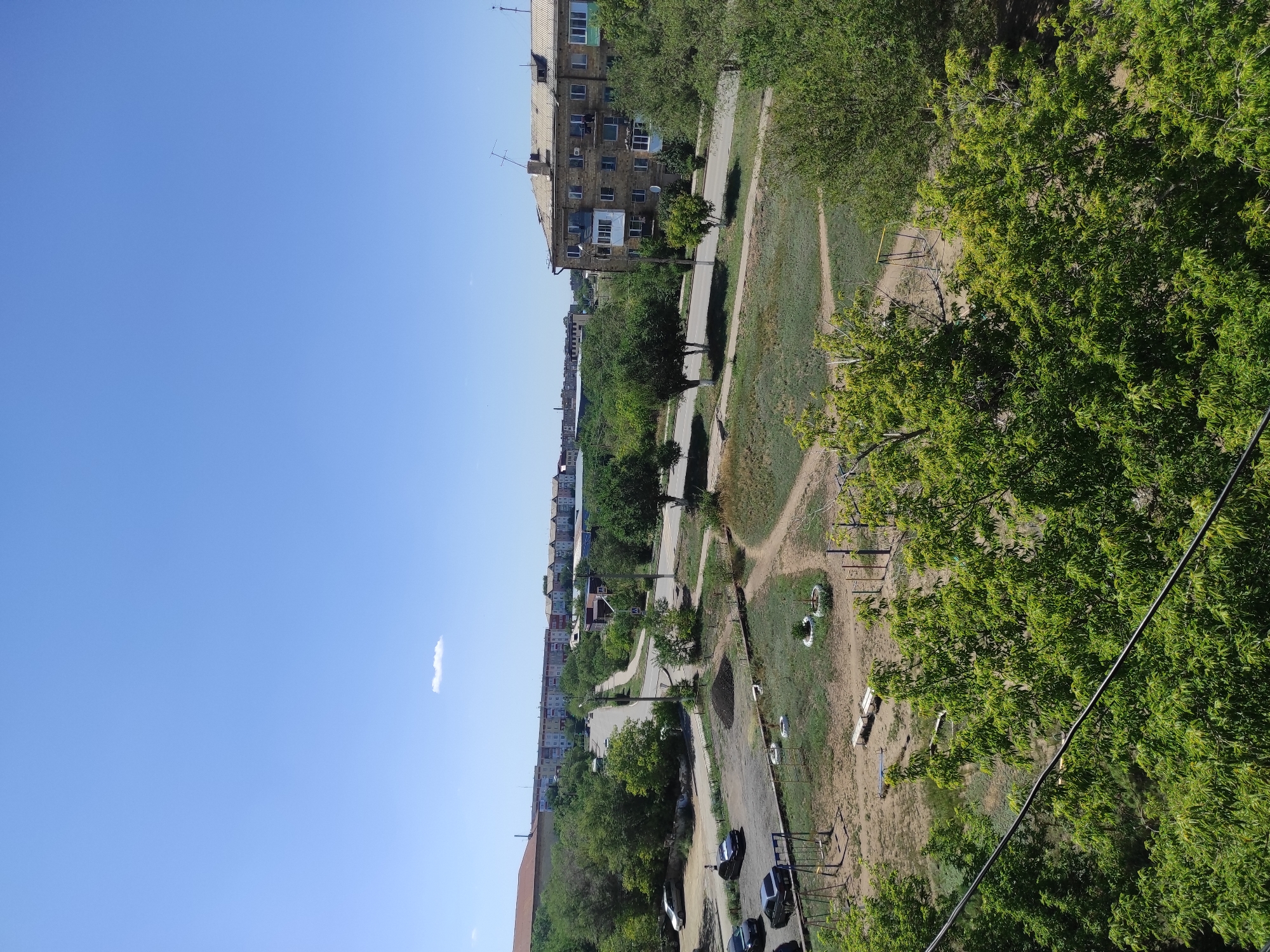 The image shows a sunny day in a residential area of Shakhtinsk, Kazakhstan. In the foreground, dense green trees with lush foliage dominate the lower portion of the frame, with a power line cutting diagonally across. Beyond the trees, a dusty playground area is visible, featuring swings and some benches. A pile of dark soil or ash is present in the middle ground, adjacent to a paved area where several cars are parked. A road winds through the scene, with more trees and vegetation lining its edges. In the background, a row of multi-story apartment buildings, typical of Soviet-era architecture, stretches across the horizon. The sky is a clear, bright blue with a single, small white cloud. The overall impression is of a typical urban residential neighborhood during daytime.