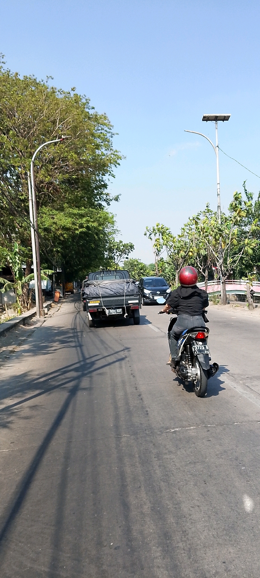A vertical, eye-level, medium shot captures a street scene under a clear blue sky in Bekasi, Indonesia. In the foreground, a person wearing a black jacket, grey pants, and a red helmet rides a black motorcycle. They are positioned in the right lane of a paved road. Behind them, a dark-colored pickup truck, also in the right lane, is covered with a black tarp secured by ropes. The truck's license plate is visible, reading "B 5590 KAV". To the left of the truck, a black car is in the left lane. The road is lined with trees and vegetation on both sides, and a lamppost with a solar panel is visible on the right. Shadows of trees and possibly other objects are cast on the road surface. The overall impression is that of a bright, sunny day with moderate traffic.