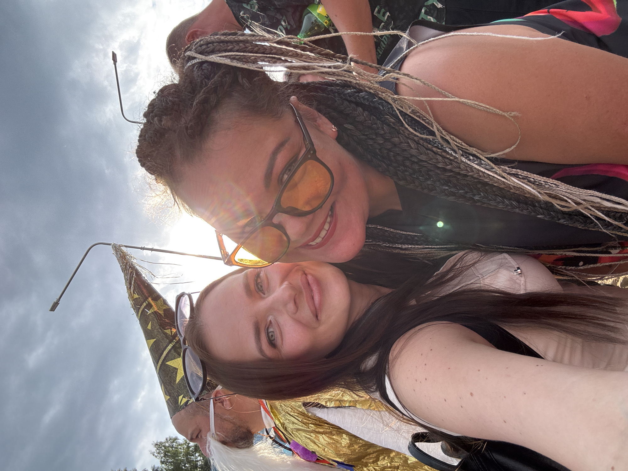 A selfie is taken outdoors in Chaikovsky, Russia, featuring two women. The woman on the left has long brown hair and is wearing a light-colored top and a black backpack. The woman on the right has braided hair and is wearing sunglasses and a black top. Behind them, a man in a golden costume and a wizard hat is visible, along with a streetlight and a cloudy sky. The overall scene appears to be festive, possibly at an outdoor event or celebration.