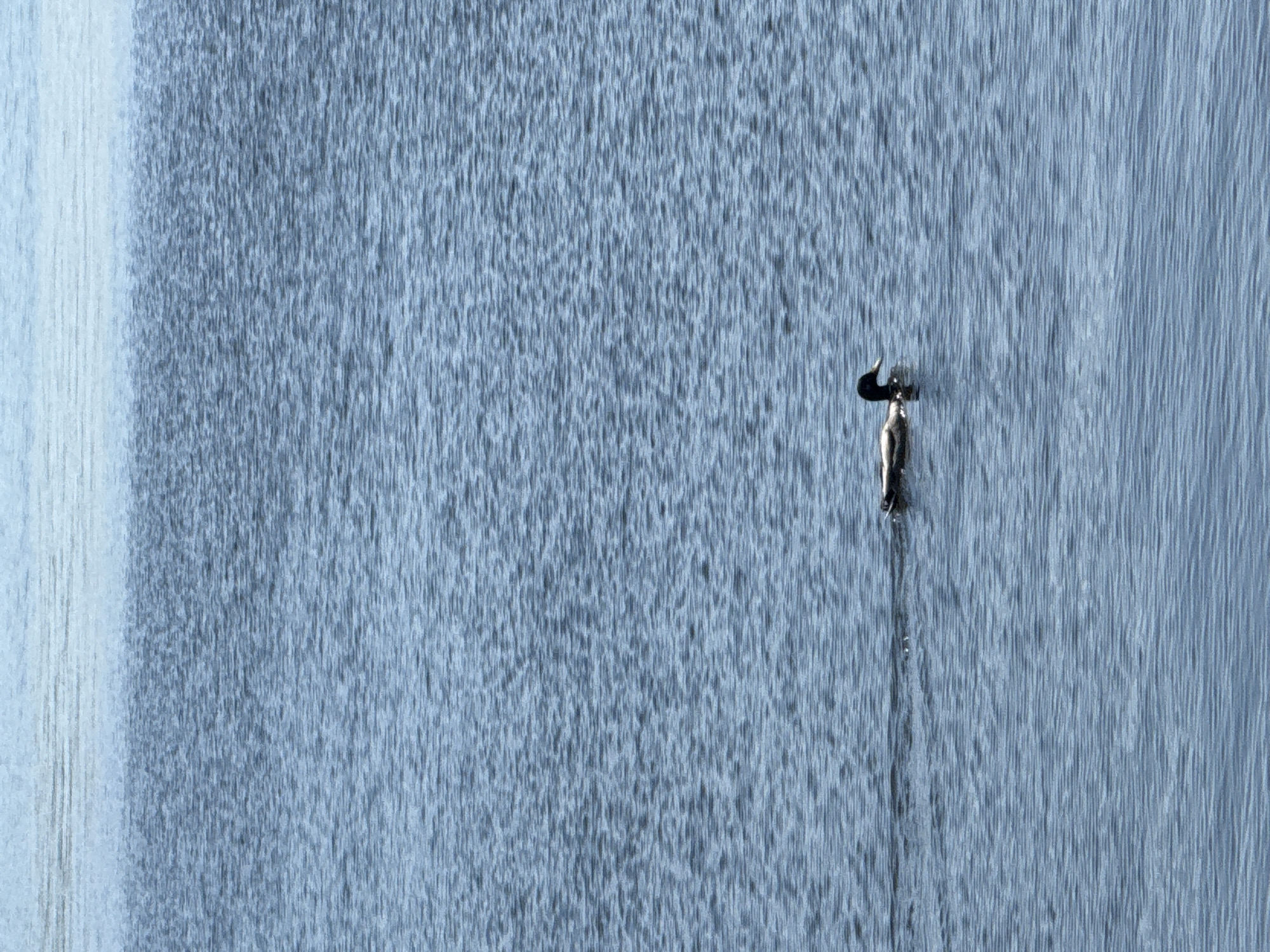 A close-up, long-shot depicts a cascade of water flowing down a wall with a metallic sculpture of a person clinging to it. The cascade appears to be a man-made water feature, and its smooth flow creates a textured pattern. To the left, the water flows more smoothly. Positioned towards the right side of the image, the sculpture is small in scale. Water streams down the sculpture, creating a visual connection with the cascading water. The image is monochromatic, primarily in shades of gray and blue. The location is in an unknown location.