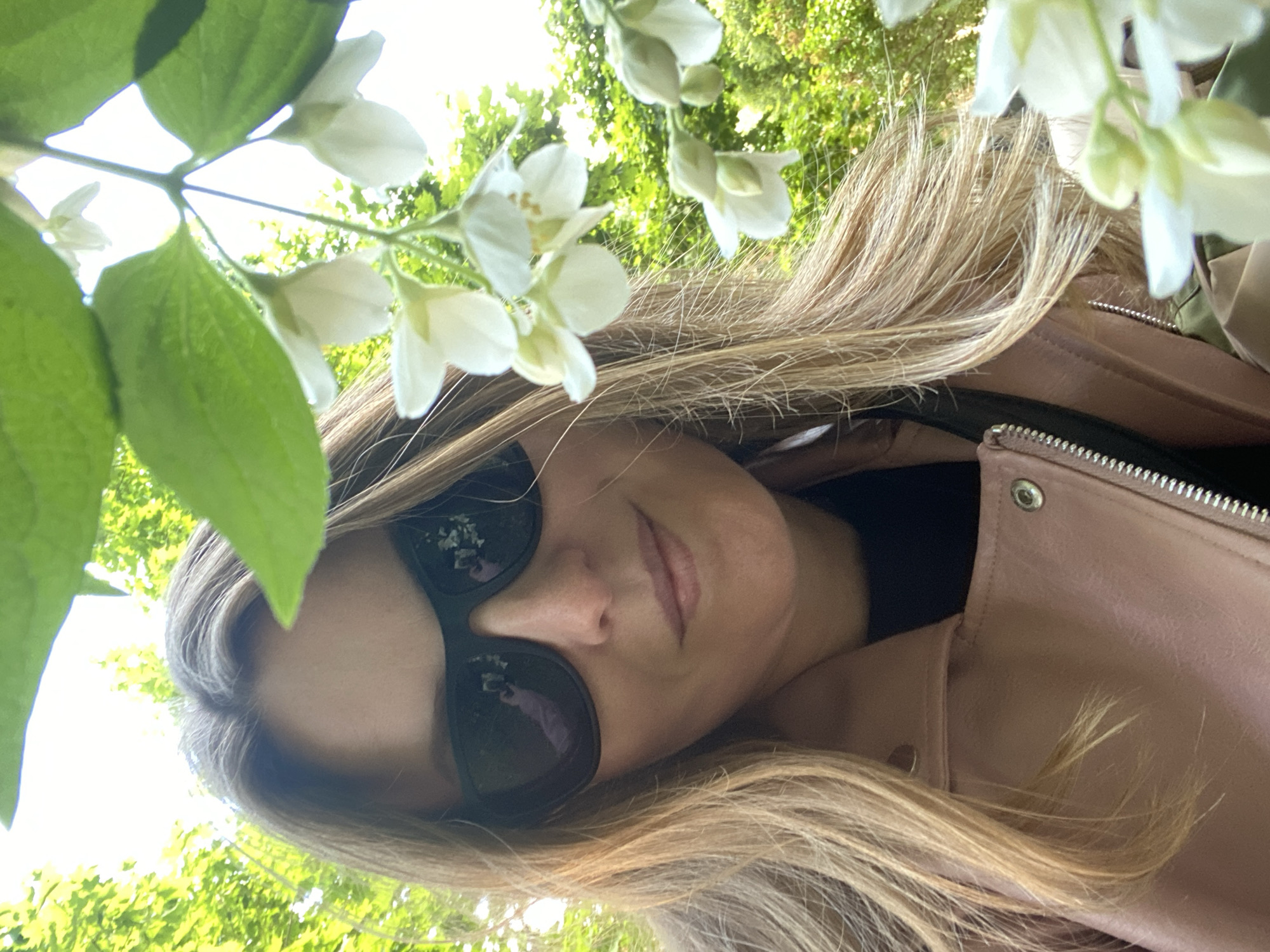 A woman with blonde hair and sunglasses, wearing a pink jacket, is the subject of the image. The background features green foliage and white flowers, suggesting an outdoor setting. The location cannot be determined from the image.