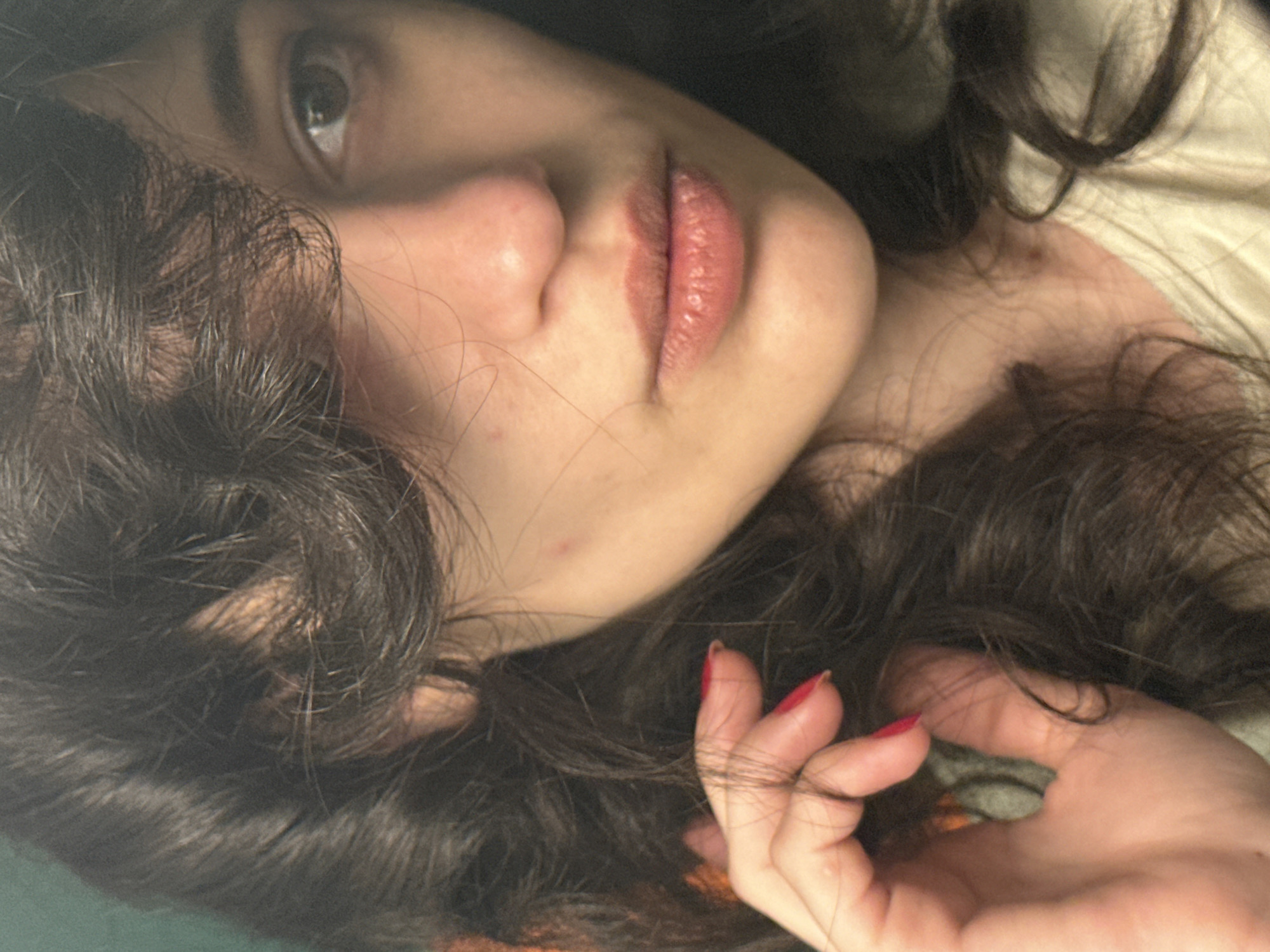 A close-up, upper-body portrait of a young woman with dark, wavy hair partially obscuring her face. Her right hand, with bright red nail polish, is raised and touching her hair near her temple. Her left eye is visible, looking towards the camera. Her lips are slightly parted, revealing a subtle lip color. The background is a muted green. There is no indication of location in Sofia, Bulgaria.