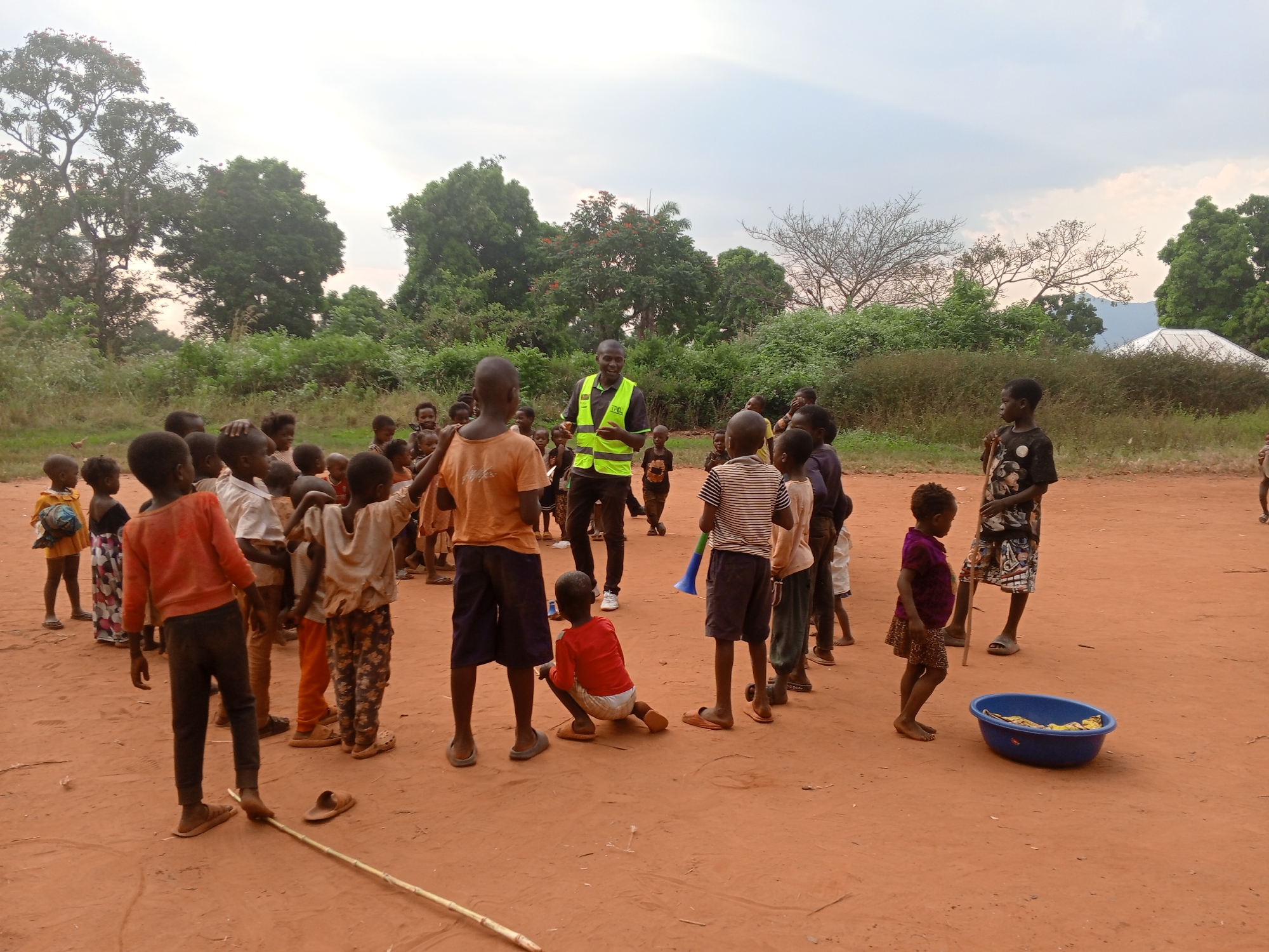 In Ngalula, Democratic Republic of the Congo, a group of children gathers on a reddish-brown dirt ground. An adult male stands among them, wearing dark clothing and a bright yellow vest with the letters "IPC" visible. The children are dressed in various casual clothes.
The environment includes green trees and shrubs in the background. A blue basin sits on the ground near some of the children.