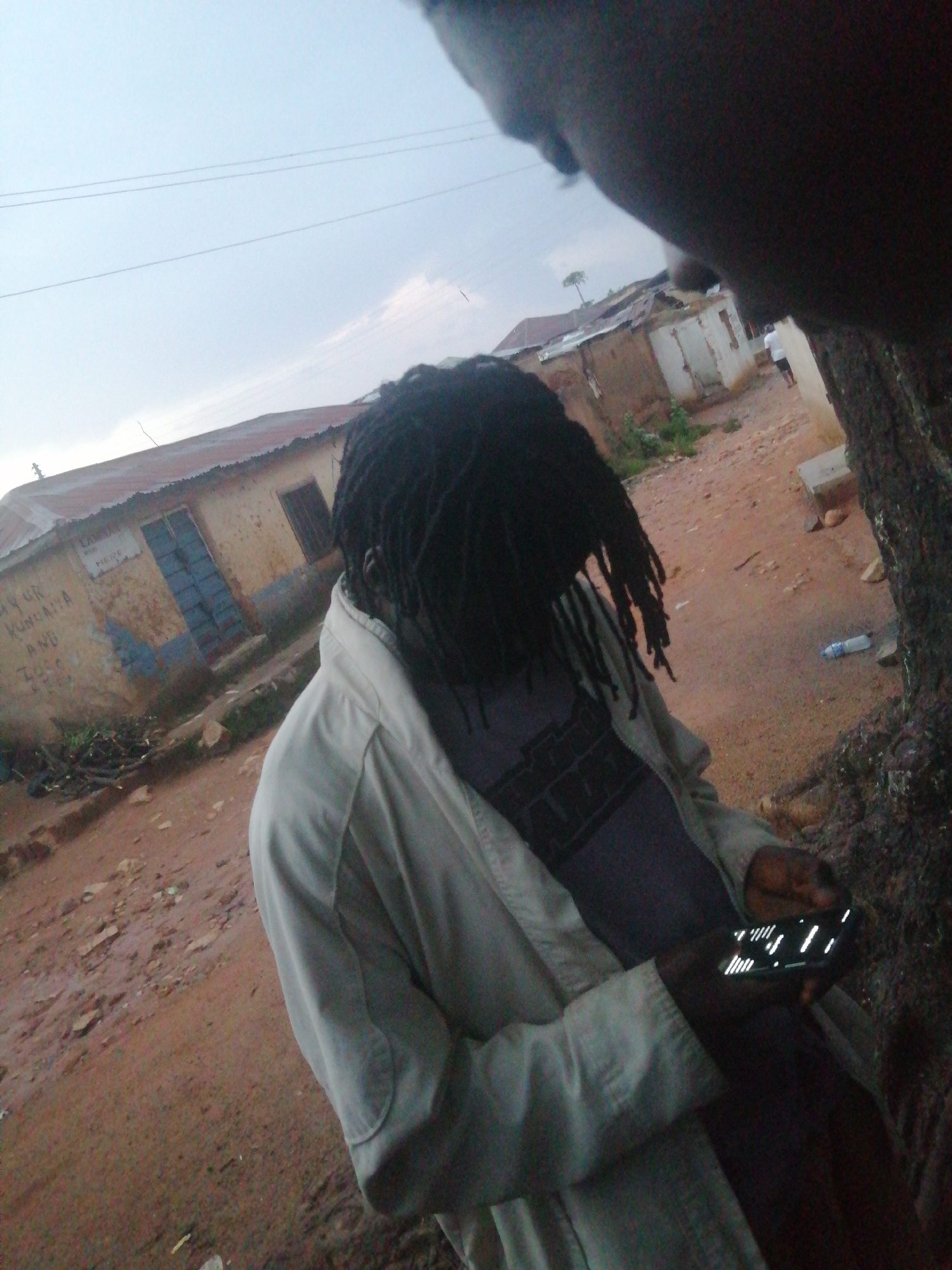 The image shows a person with dreadlocks wearing a light-colored jacket, holding a smartphone. The person is in an outdoor, likely urban, environment with buildings and dirt roads visible in the background. The buildings appear to be made of brick and corrugated metal, with some showing signs of wear and tear. There are also some power lines overhead. The ground is a mix of dirt and gravel, with some scattered debris. The lighting suggests it is daytime, possibly overcast. The focus is on the person and their interaction with the smartphone.