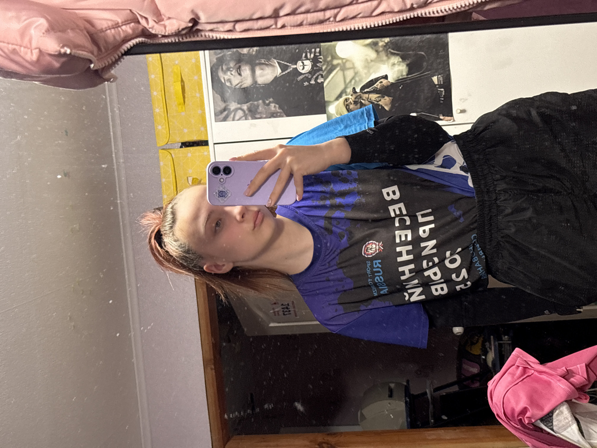 A young person, likely female, is captured in a mirror selfie in an indoor setting, possibly a bedroom in Прикамский, Russia. The image is rotated 90 degrees counter-clockwise.
The subject, with light brown or auburn hair pulled back, stands with a neutral expression, holding a purple smartphone in her right hand to take the photo. Her fingernails are painted in a light color. She wears a blue long-sleeved shirt underneath a purple and black short-sleeved top, which features white and blue text and designs. Black athletic shorts or pants complete her attire.
The reflected background reveals parts of a room. Directly behind the subject's head, two black and white framed pictures or posters are visible on a light-colored wall. The left poster depicts a person wearing a prominent necklace with a cross-like pendant. The right poster shows another person or a scene. To the left of these posters, a stack of yellow storage boxes or drawers with a subtle pattern is reflected. The mirror itself has some visible smudges or dust. In the upper left corner of the reflection, part of a pink puffy jacket hangs. In the lower right, a crumpled pink garment and other clothes are visible.
Visible text on the subject's top, reflected in the mirror, includes Cyrillic letters that appear as "ВЕСЕННИЙ" (Vesenny - Spring) and "СПОРТ" (Sport), along with other partially legible words and "CONB...". Some small text like "J431" and "TEAC" is also faintly visible on a yellow object in the background.