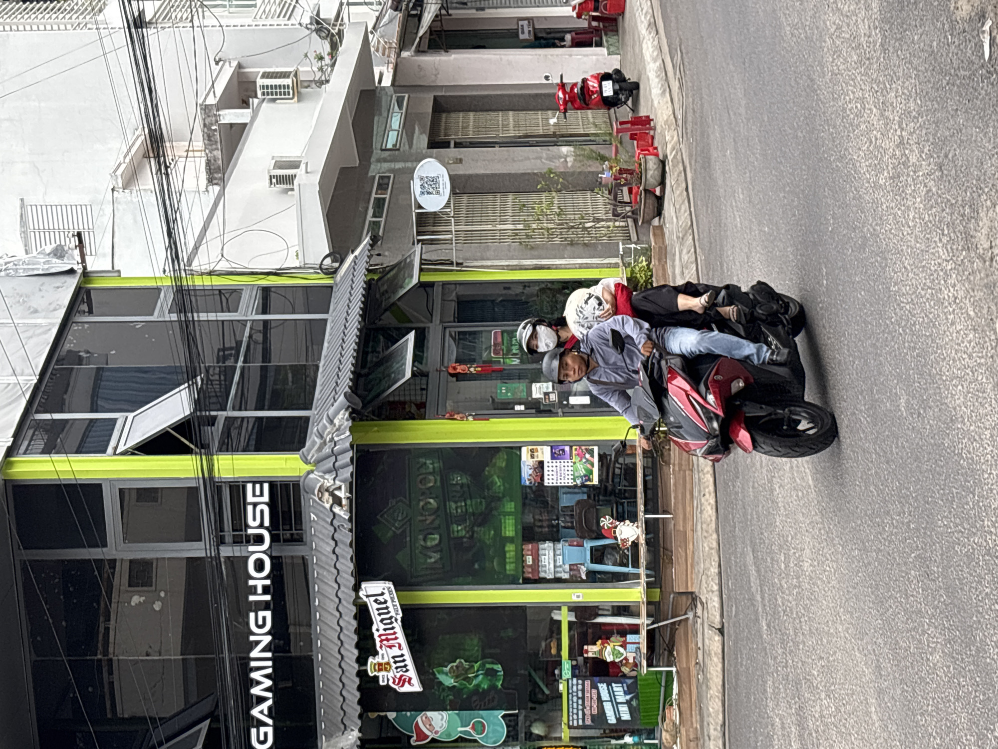 In Nha Trang, Vietnam, a red and black motorcycle carrying two individuals, a driver wearing a grey jacket and blue jeans, and a passenger wearing a white shirt and dark pants, travels on a paved road. The driver wears a black helmet, and the passenger wears a white helmet and a face mask. The motorcycle's front is visible. In the background, a building with a glass facade displays a neon sign that reads "GAMING HOUSE" and "MINI MART". Next to it, a store with a green facade exhibits a San Miguel beer advertisement and other merchandise. The street features several potted plants and red plastic stools outside another establishment. The weather appears overcast, with a slight haze in the air.