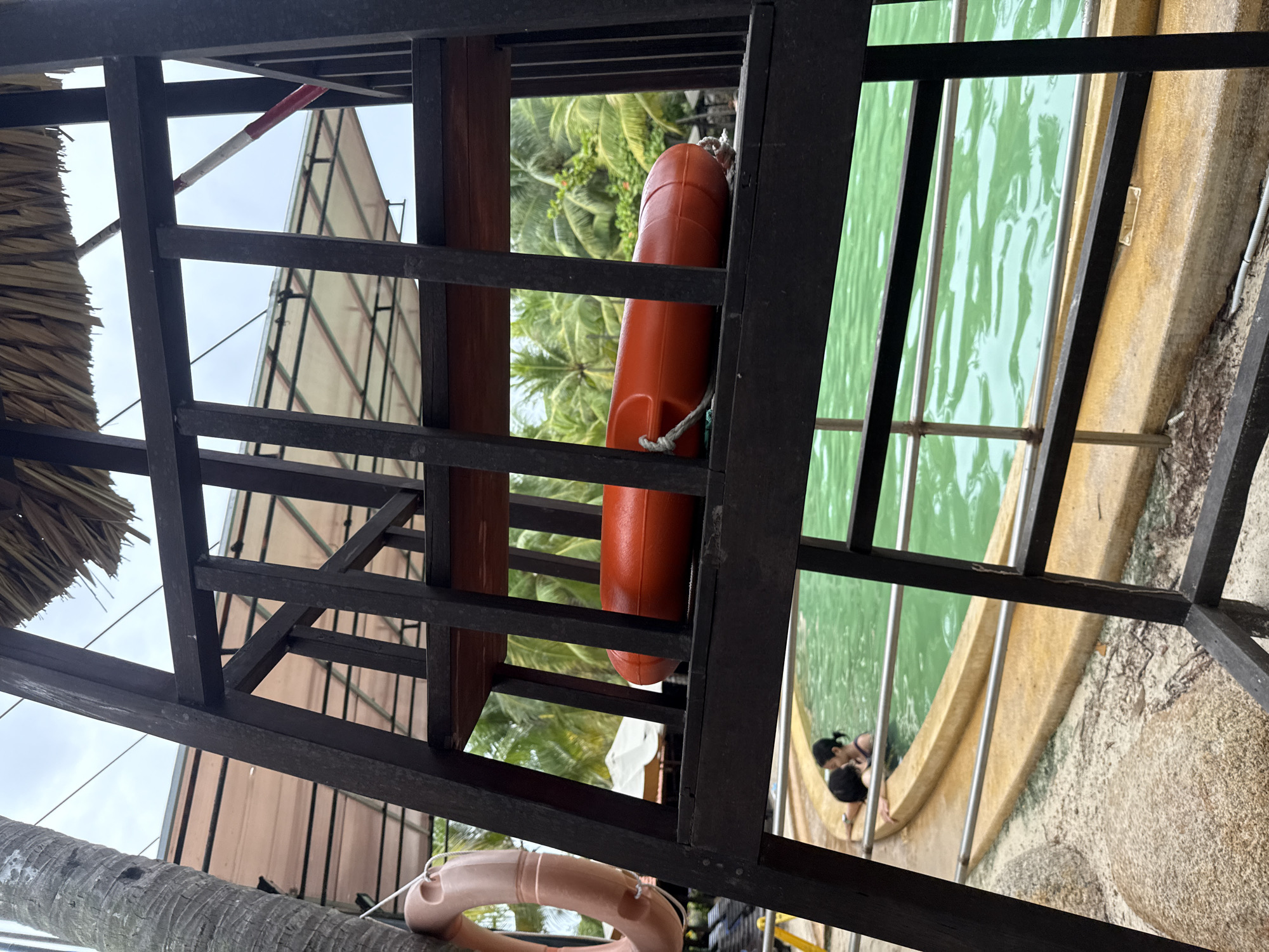 The image depicts a lifeguard chair at a swimming pool area, likely in Nha Trang, Vietnam, characterized by lush palm trees in the background. The chair is constructed of dark wood with horizontal slats. On the second shelf of the chair, an orange flotation device with a rope attached is visible. To the left of the chair, partially obscured by the palm tree, a tan-colored life preserver ring is hanging. In the background, a person is partially submerged in the bright green water of the swimming pool, suggesting a resort or public swimming facility. The overall setting is tropical, with a cloudy sky overhead.