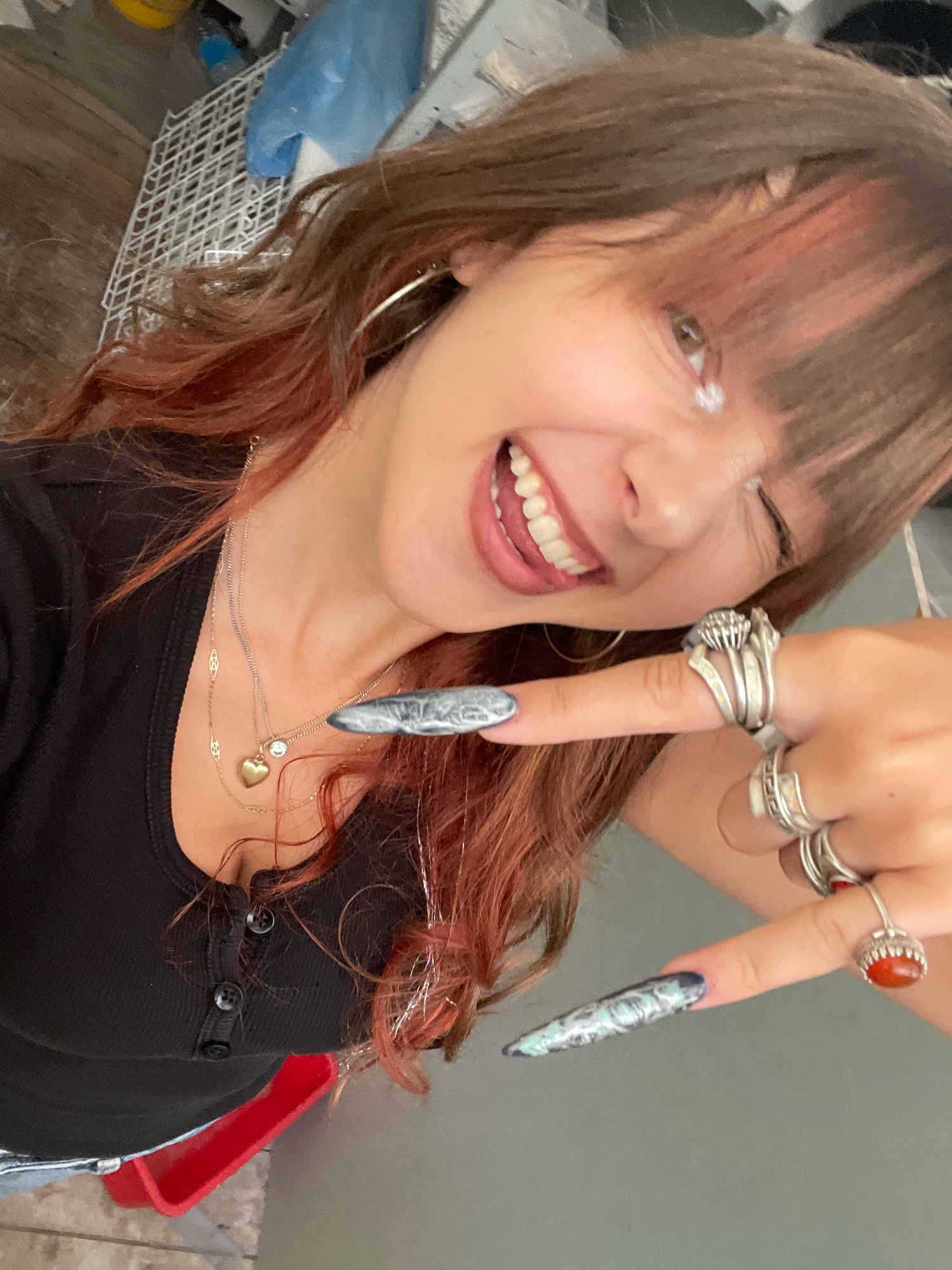 A young woman with reddish-brown hair and bangs is taking a selfie indoors. She is wearing a black henley shirt and multiple silver rings on her fingers, one with a red stone and another with a unique abstract design. Her fingernails are long and painted with a marbled pattern. She is smiling and winking, with glitter applied to her eyelids. She is wearing a delicate gold necklace with a heart-shaped pendant and a small diamond. The background is blurred and shows what appears to be a cluttered area with a white shelf unit, a blue plastic bag, and wooden flooring. The image does not provide any specific information or context related to the city of Raanana, Israel.