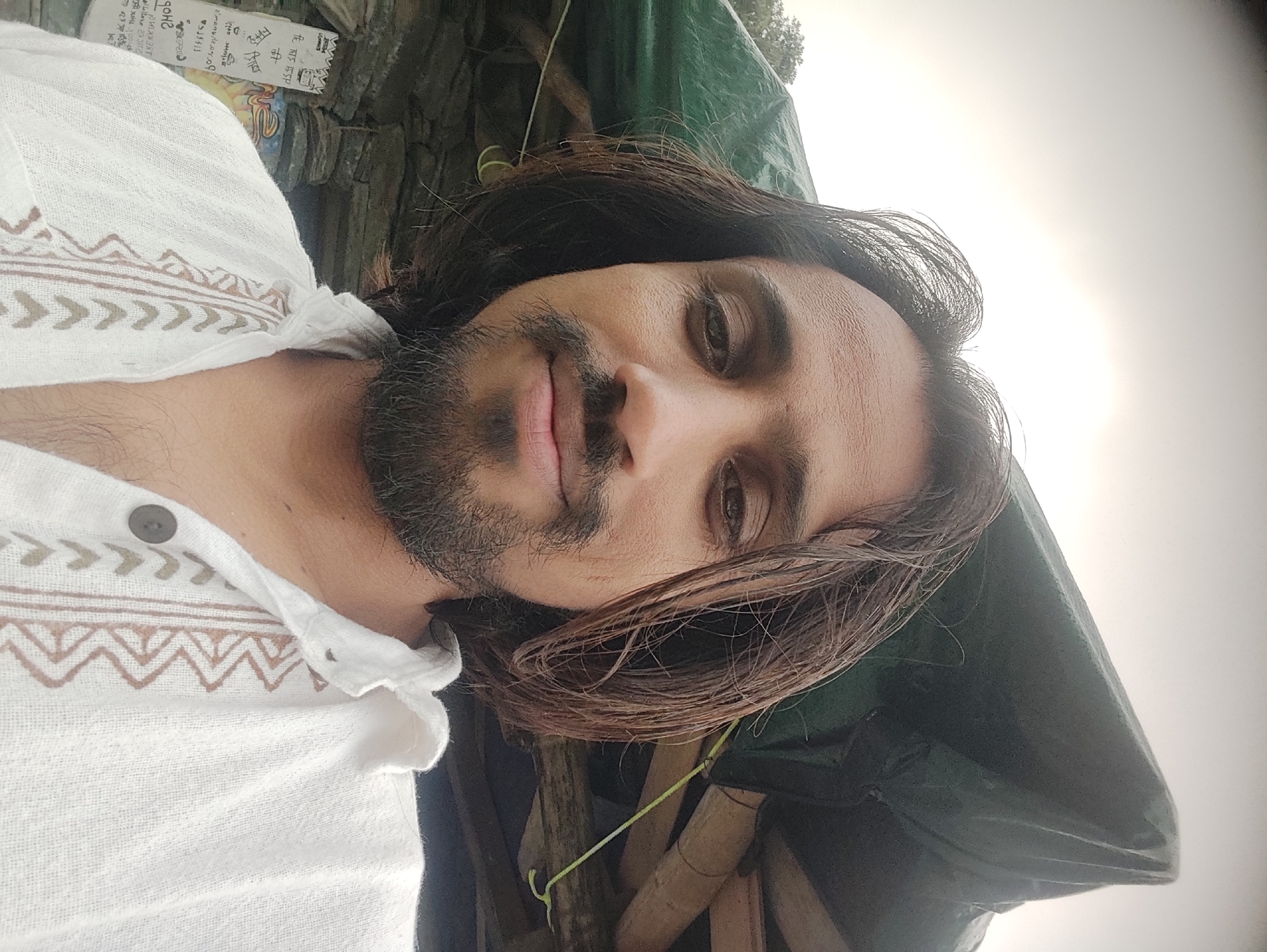 A close-up, eye-level selfie shows a man with shoulder-length dark hair and a beard, looking directly at the camera. He is wearing a white, embroidered shirt. Behind him, there is a dark green tarp partially covering some wooden structures and what appears to be a stone wall. The sky above is overcast. The scene is likely outdoors, possibly in an area with rustic shelters, and the mention of Bhagsu, India, suggests a location in a hilly or mountainous region.