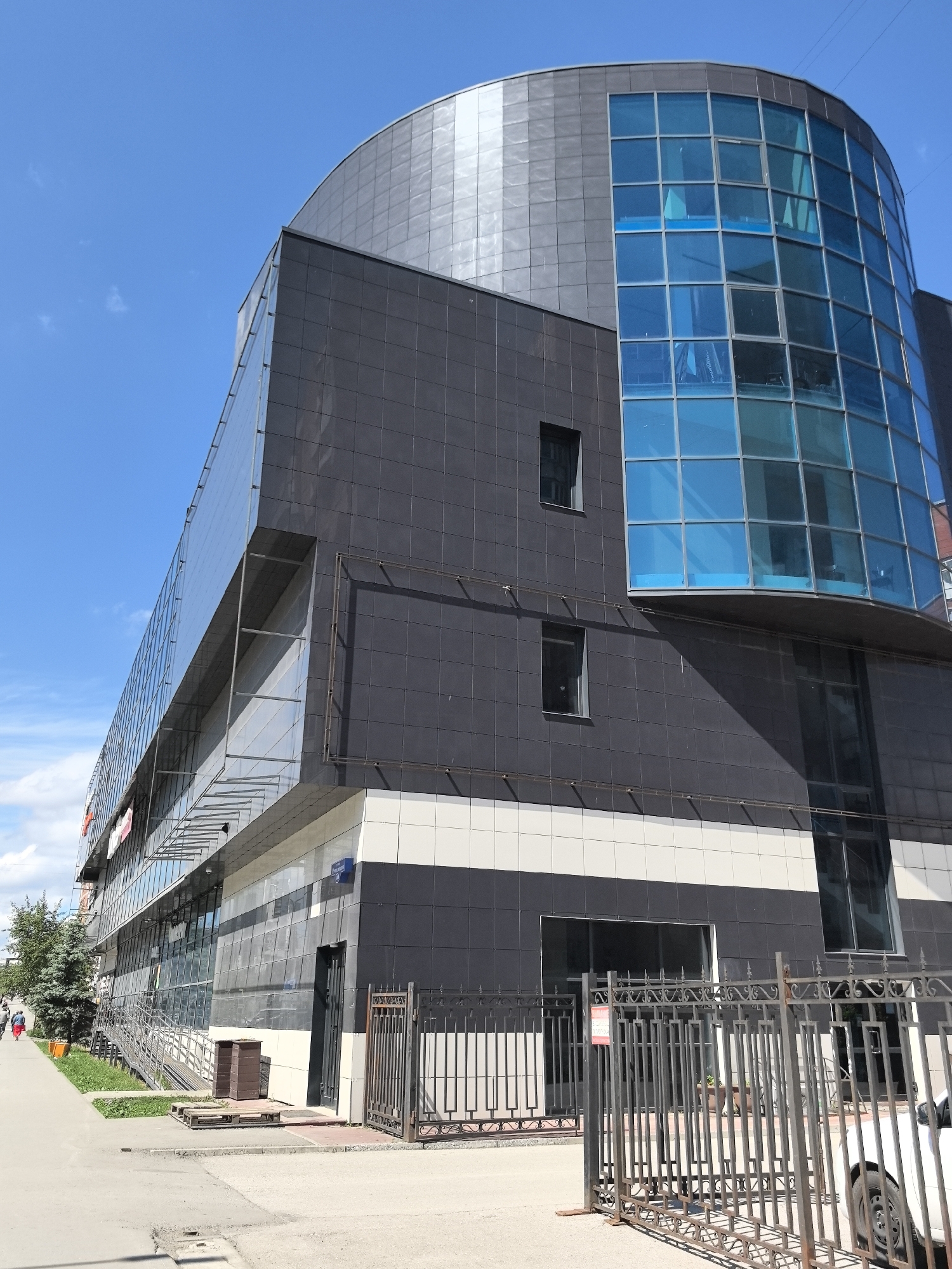 The image shows the exterior of a modern building in Perm, Russia. The building features a combination of architectural styles, with a curved, metal-paneled roof section, a large area of blue-tinted windows, and a lower section clad in dark gray and white rectangular panels. The structure has multiple stories, with at least two floors visible from this angle. A metal fence runs along the sidewalk in front of the building, partially obscuring the view of the ground floor. Part of a white car is visible behind the fence. The sidewalk is wide and paved. In the background, a few trees and other buildings are visible. The sky is bright blue with scattered clouds. A person in red clothing is walking on the sidewalk in the distance.