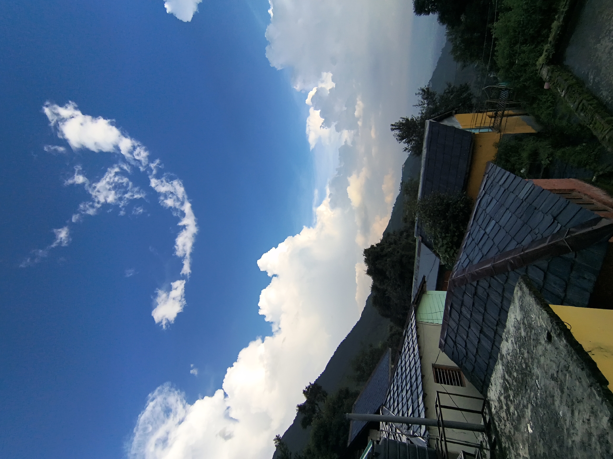 The image depicts a village scene in the mountains, likely in a region like McLeod Ganj near Dharamshala, Himachal Pradesh, India, which is a hub for Tibetan Buddhism and has a significant population of Tibetan refugees. The village is situated on a hillside, with houses built on terraced slopes. The houses have dark, sloped roofs, typical of the region's architecture. The foreground shows the weathered concrete roof of a building and the tiled roofs of several houses. In the background, a steep, tree-covered mountain slopes upwards, disappearing into the sky. The sky is a vibrant blue with dramatic, fluffy white clouds, suggesting fair weather with some cloud cover. The overall impression is one of a peaceful, rural mountainous landscape.
