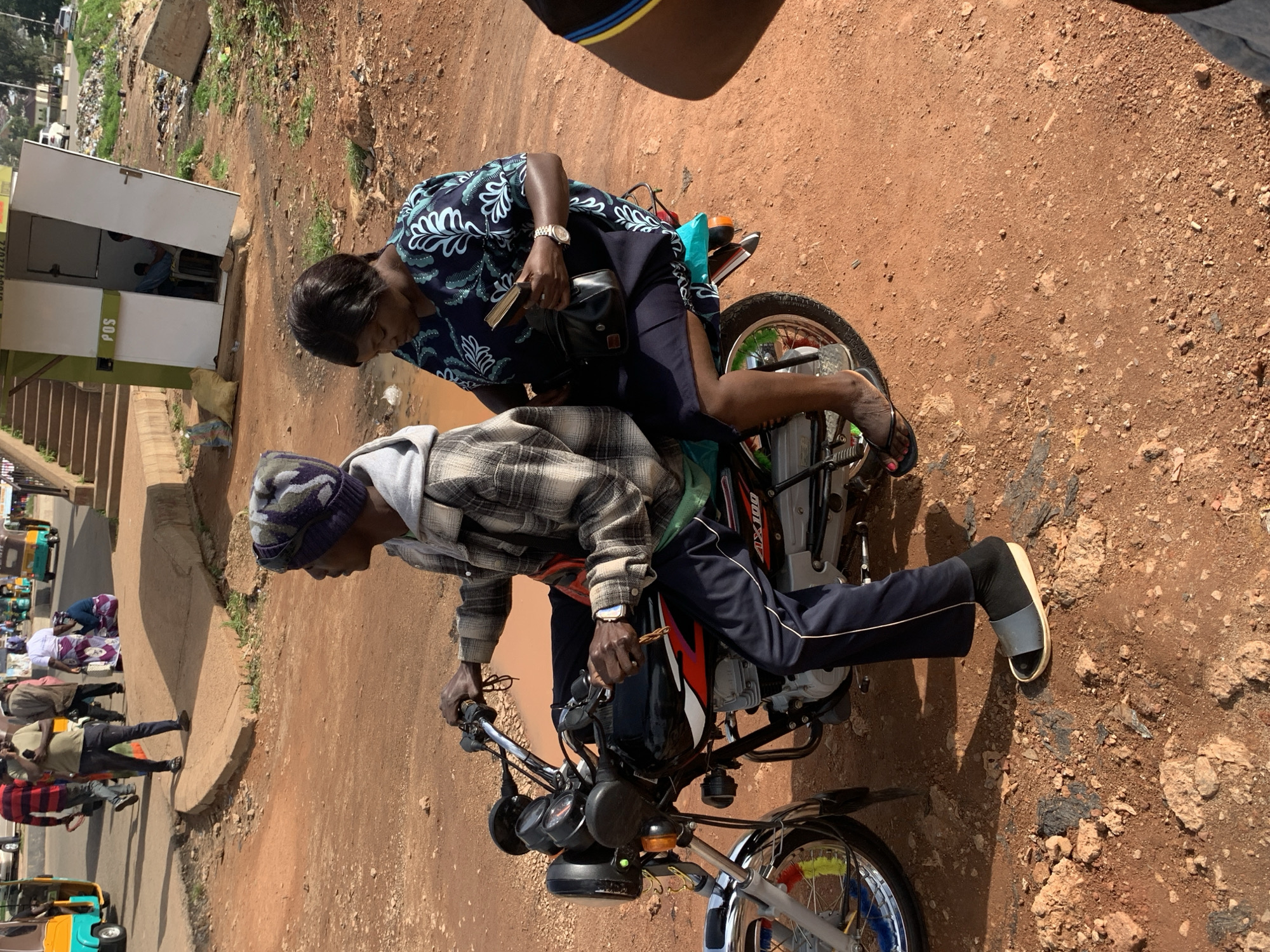 In Gura Top, Nigeria, two individuals are seated on a motorcycle on a dirt road. The driver, a man wearing a plaid shirt, a grey hooded jacket, and a beanie, is holding the handlebars. A woman, dressed in a dark blue patterned dress, is seated behind him, holding a book in her right hand. The motorcycle is black with red and white accents and the model "AX100" is visible. In the background, a small white building with an open door, labeled "POS," stands near a staircase. Several vehicles, including auto-rickshaws, are visible further in the distance, along with scattered debris. The ground is a mix of reddish-brown dirt and rocks.