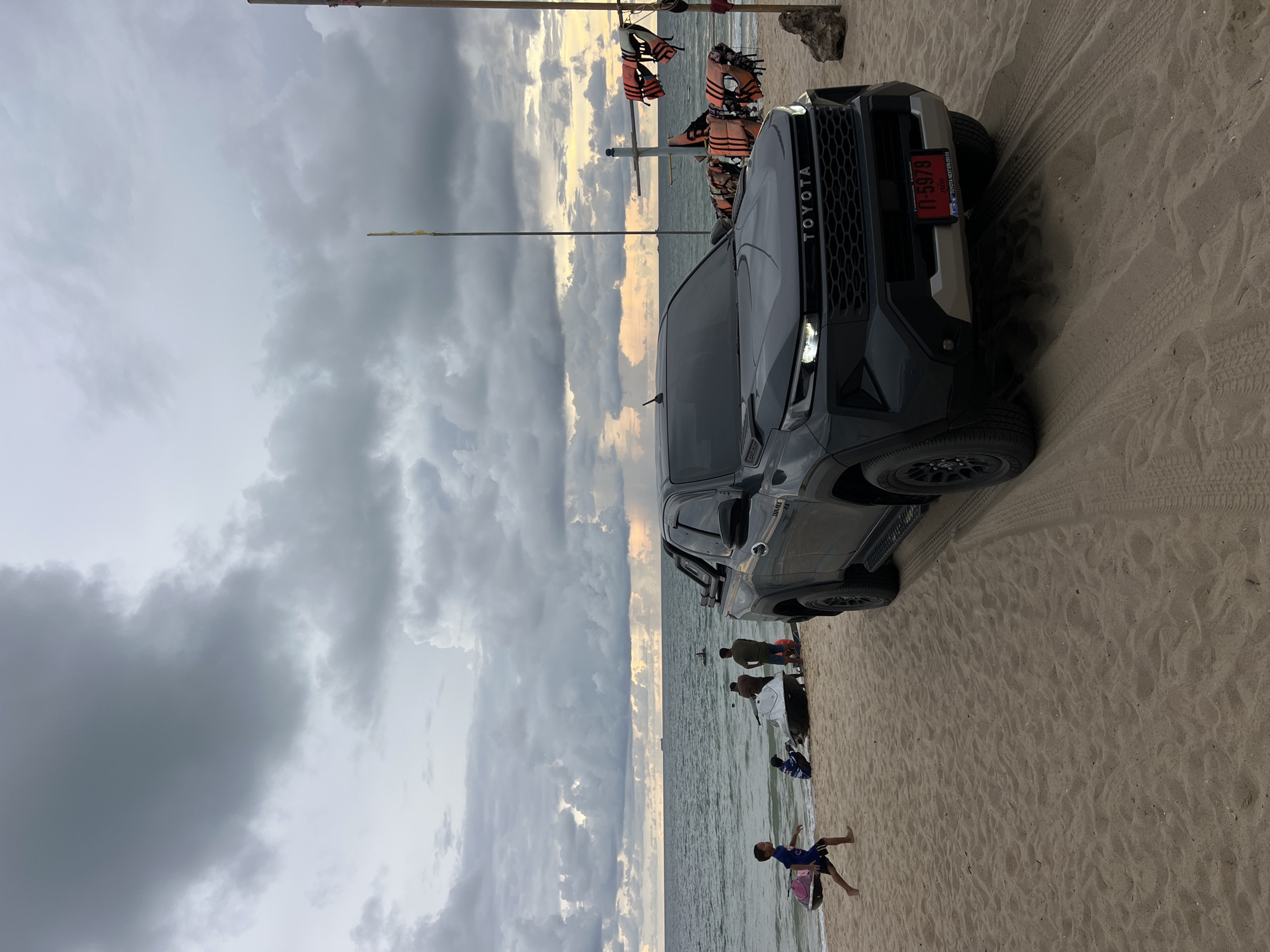 A dark gray Toyota Fortuner SUV is parked on a sandy beach next to the ocean. The vehicle is angled towards the water, with its front facing the viewer. The sky is overcast with dark gray clouds, but there is a hint of yellow and orange light peeking through on the horizon, suggesting either sunrise or sunset. Several life jackets are hanging on a pole behind the SUV.
A few people are visible on the beach and in the shallow water. In the foreground, a young child in a blue shirt and dark shorts is standing and appears to be playing. Further back, several other individuals are gathered near the water's edge, some appearing to be looking at the ocean or interacting with each other. The beach is covered in sand, and tire tracks are visible behind the SUV, indicating it has driven on the sand.
The overall scene suggests a casual day at the beach, possibly in the late afternoon or early morning given the lighting. The weather appears to be changing, with the clouds hinting at potential rain or a dramatic sky. Visible text on the license plate of the SUV reads "กท 5978". The word "TOYOTA" is clearly visible on the front grille of the vehicle.