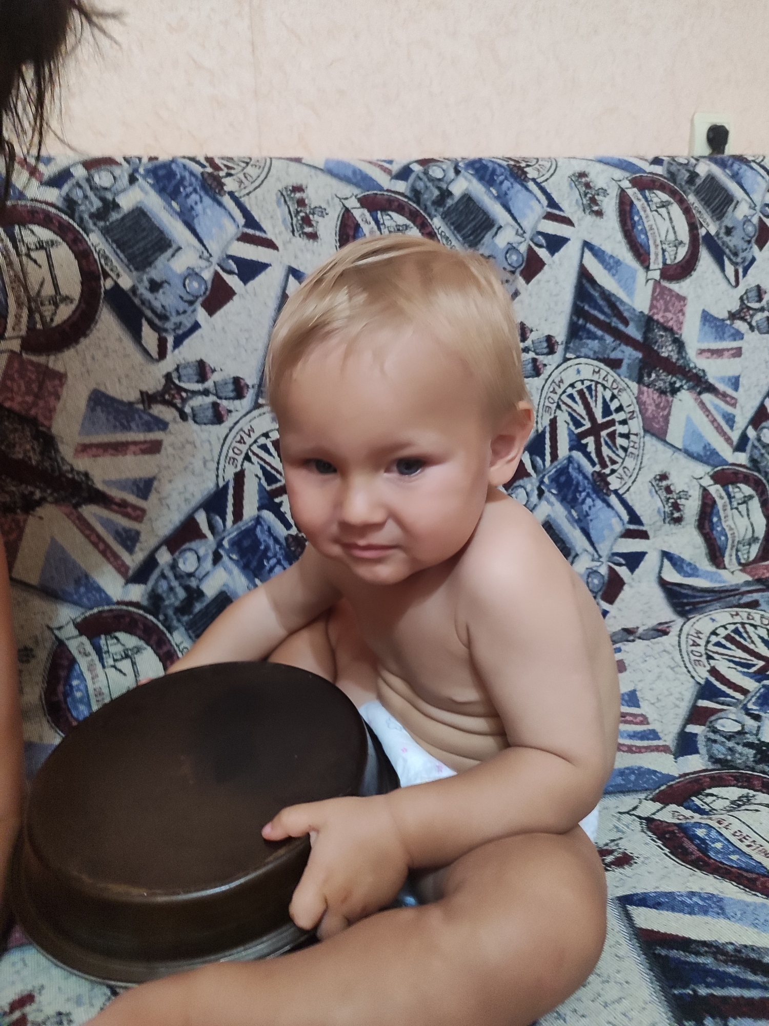 A close-up, medium shot captures a young child sitting on a sofa with a patterned blanket. The child, with fair hair and blue eyes, is wearing a diaper and looking towards the right side of the frame with a slight pout. Their hands are resting on a dark, circular object, possibly a pot lid. The sofa's blanket features a repeating pattern that includes images of London buses, the Union Jack flag, and circular emblems with text. The background appears to be a wall, lightly textured and a pale peach color. The lighting suggests it is daytime, with no indication of specific weather conditions. The overall mood is informal and domestic, focusing on the child's interaction with the object and their expression. Visible text on the blanket includes "MADE IN THE UK" and "LONDON". The scene is set in Tuapse, Russia.