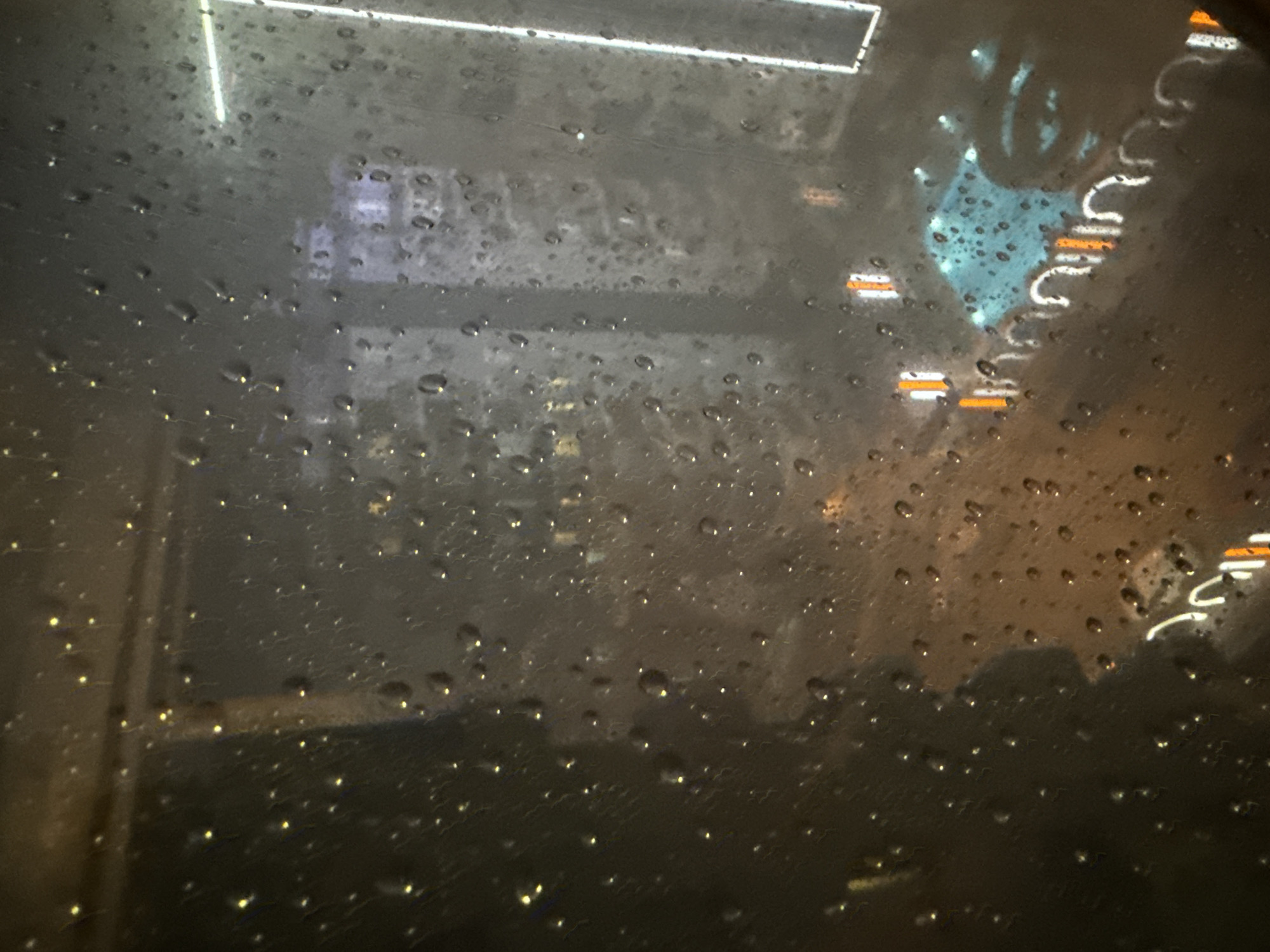 A rainy window obscures a night scene. Light reflects on droplets. In the distance, building lights and streetlights indicate an urban environment. A large glowing sign reflects on the upper right of the window.
