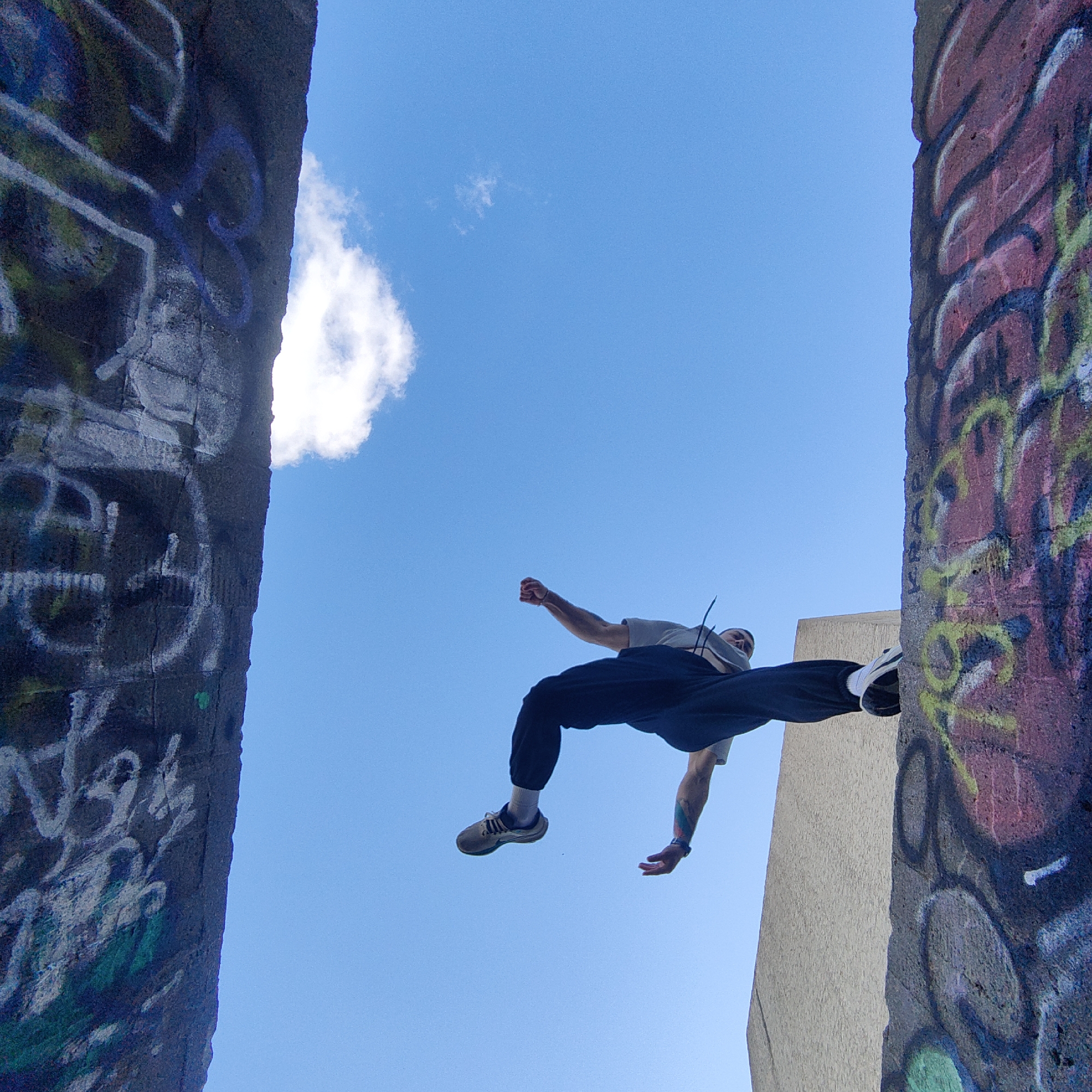 An individual is captured mid-jump between two graffiti-covered concrete structures in Varna, Bulgaria. The sky is blue with a single cloud visible behind him. The subject wears a grey t-shirt, dark pants, white socks, and athletic shoes. Graffiti covers the concrete structures on either side of the jumper.