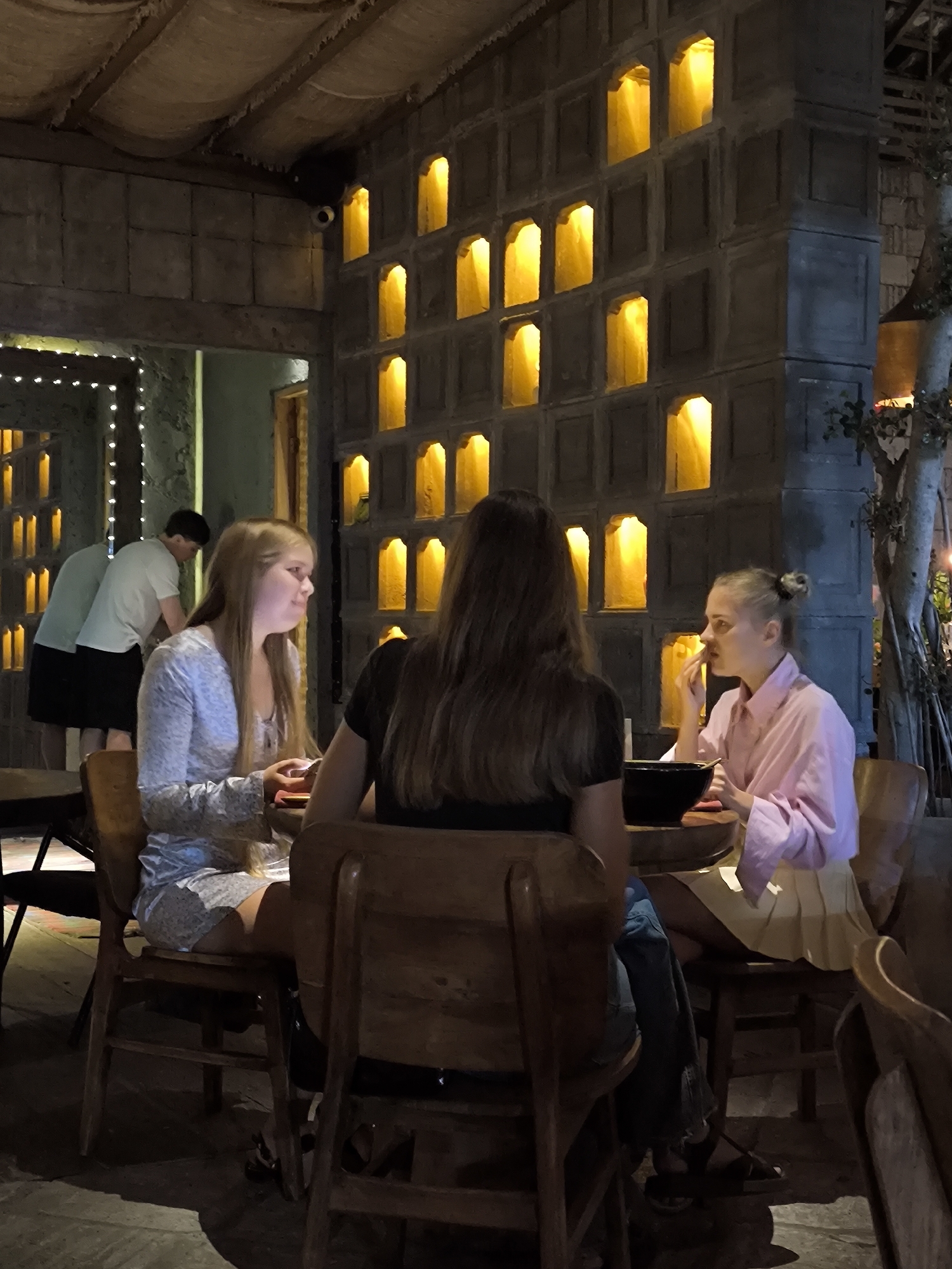 The image depicts an indoor dining or lounge area, likely in North Kuta, Indonesia, during the evening. The space is dimly lit, with a warm, intimate atmosphere created by numerous yellow lights set into recessed niches of a distinctive wall made of square stone or concrete blocks. The ceiling has a rustic, textured fabric or woven finish.
In the foreground, three young women are seated around a wooden table. On the left, a woman with long blonde hair, wearing a light patterned top, looks towards her companions with a slight smile. Facing away from the camera, a woman with long dark hair, dressed in a dark top, sits between her two friends. On the right, a woman with blonde hair tied back, wearing a pink collared shirt and a light skirt, is engaged in eating from a dark bowl. Their chairs are made of dark wood, consistent with the rustic decor.
In the background, two other individuals are visible. On the left, a person in a light shirt and dark shorts stands near a wall decorated with string lights. Further right, another person in a light t-shirt and dark shorts is partially visible, appearing to be working or moving about. A small security camera is mounted high on the wall in the upper left. A decorative, tree-like structure with prominent roots or branches adds to the unique interior design on the far right. No text is visible in the image.
