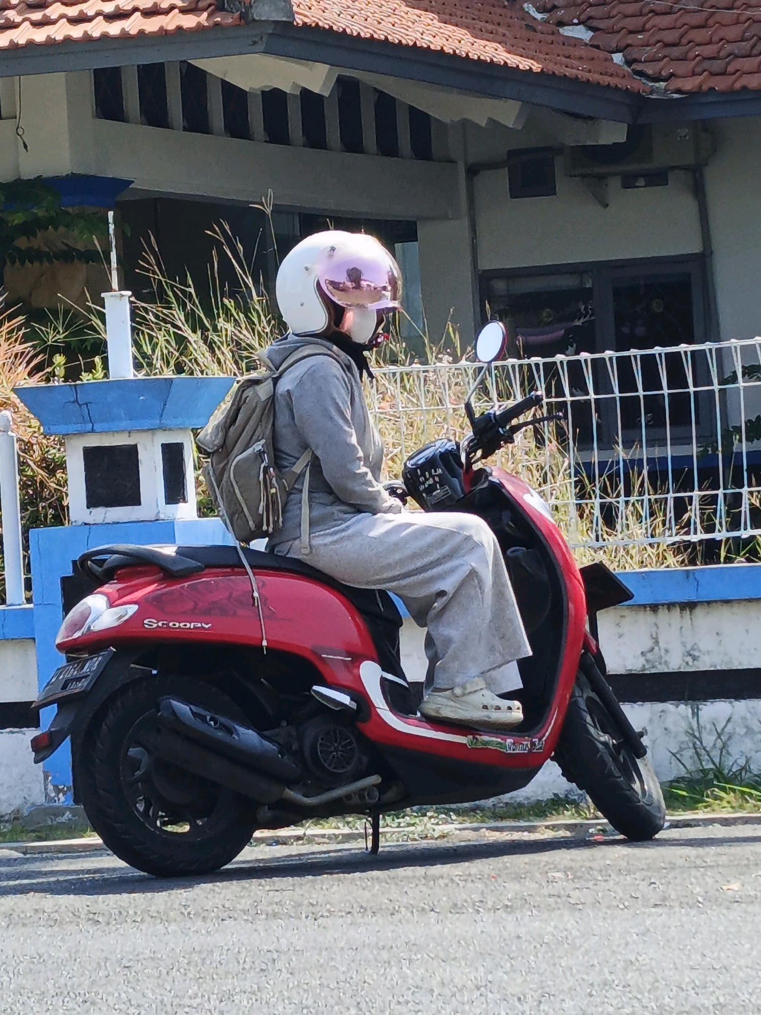 A person wearing a light gray tracksuit and a white helmet with a pink visor is seated on a red Honda Scoopy scooter. The scooter has "Scoopy" written on its side. The person is also wearing a light brown backpack. They are parked on an asphalt road, with a blue and white fence and some dry grass in the background. Behind the fence, a building with a red tiled roof and striped window shutters is visible. The scene appears to be outdoors during the daytime, under clear weather conditions. The image is taken from a side profile of the person and the scooter. The license plate on the scooter is partially visible and appears to have text and numbers, but they are not clearly legible. The overall impression is that of a casual, everyday scene in a residential or semi-urban area in Kludan, Indonesia.