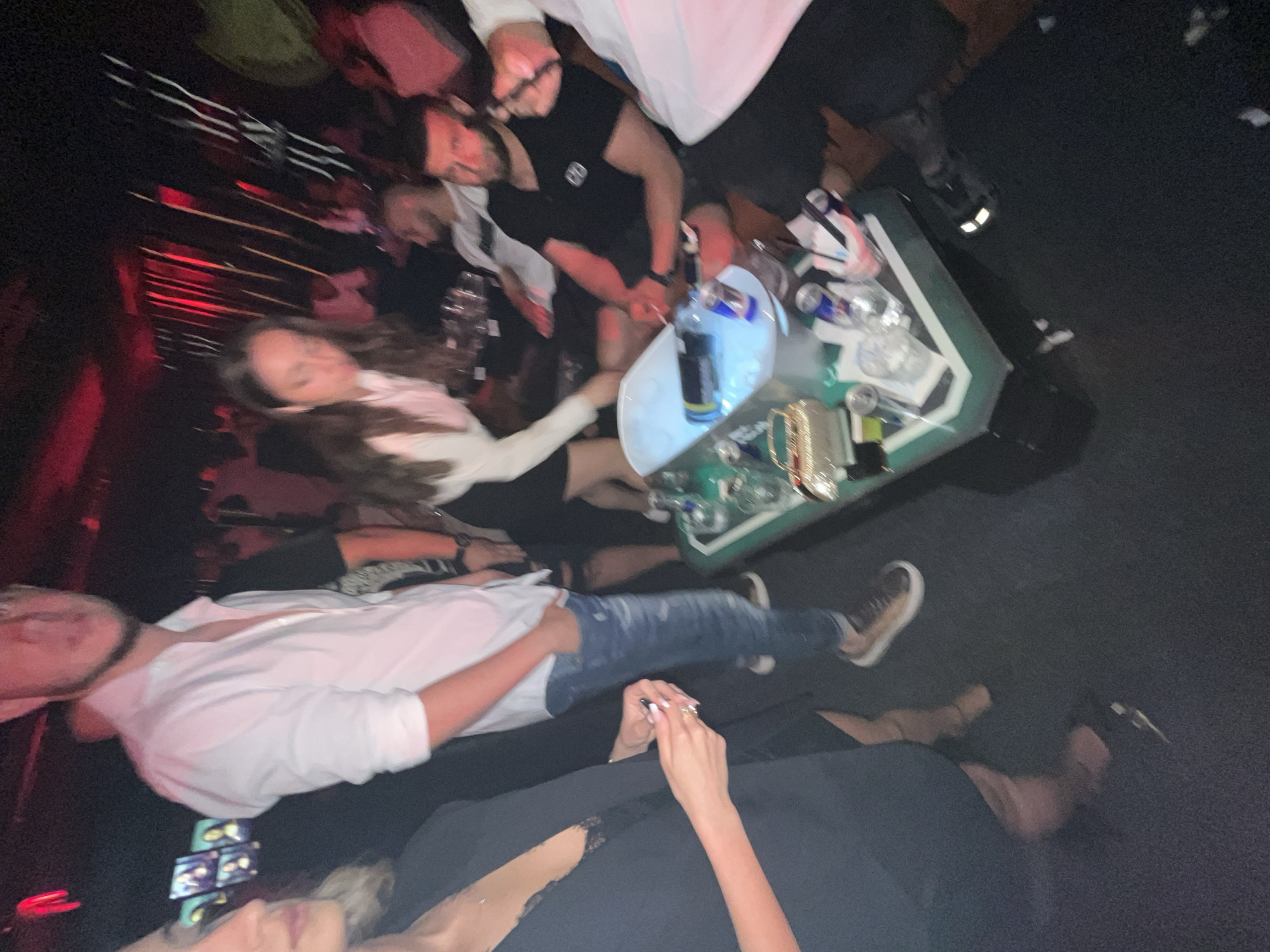 A blurred, angled-down view of a crowded indoor scene. A group of people are gathered around a table holding drinks and bottles. One person is wearing a white shirt and jeans and another is wearing a dark outfit with visible arms and hands. A green-edged table holds bottles of alcohol and what looks like a bucket of ice. Some of the people are facing the table while others are oriented in different directions. The location of the scene, as well as the city, is unidentifiable.