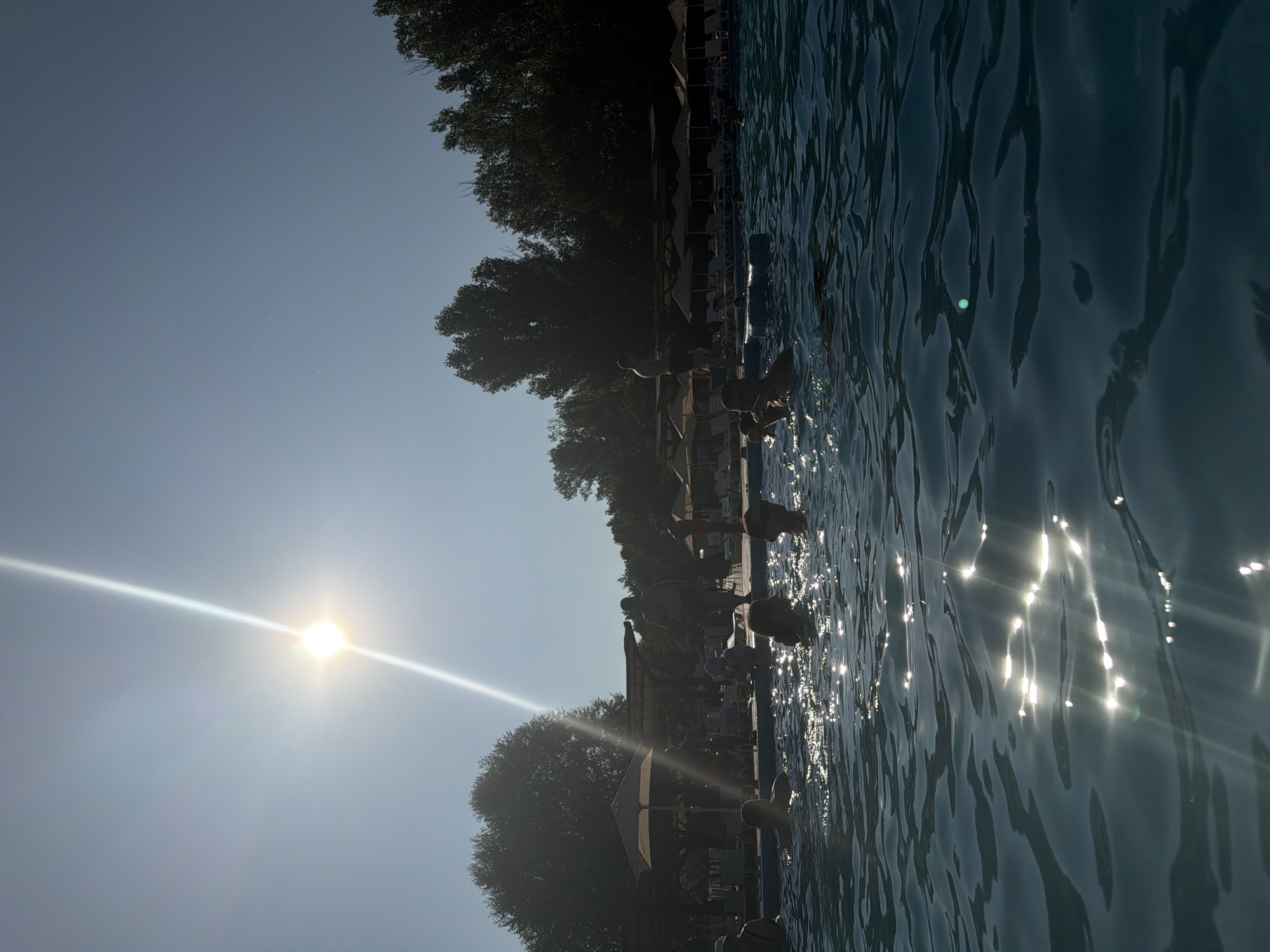 The image depicts a sunny day at an outdoor swimming pool, likely within the city of Sociteni, Moldova. The sun is high in the clear blue sky, casting a bright glare that reflects off the rippling water of the pool. Several people are visible in the water, some swimming or wading, while others appear to be relaxing. In the background, there are lounge chairs and umbrellas, suggesting a recreational area. Trees line the perimeter of the pool area. The overall scene conveys a sense of a warm summer day with people enjoying leisure activities.