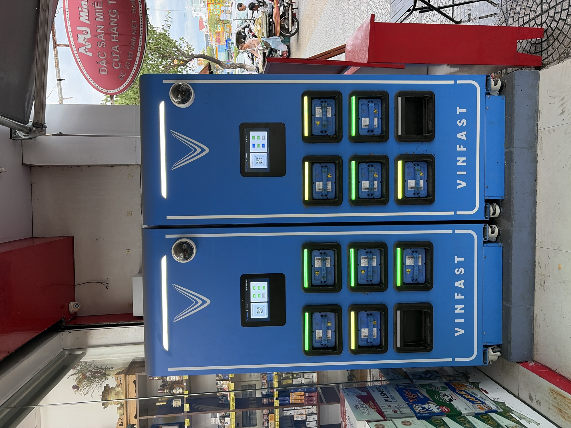 The image shows two blue VinFast battery swapping stations side-by-side on a sidewalk in Da Nang, Vietnam. The stations have screens displaying QR codes and charging status indicators. Each station has multiple compartments, some with green or yellow lights, indicating active charging or readiness. A red cabinet is located to the right of the stations, and a table and chairs are partially visible. In the background, people are seen interacting with a street vendor selling durian, and vehicles are present on the street. A sign with Vietnamese text, "ĐẶC SẢN MIỀN...", is visible above the stations. The environment appears to be an urban commercial area.