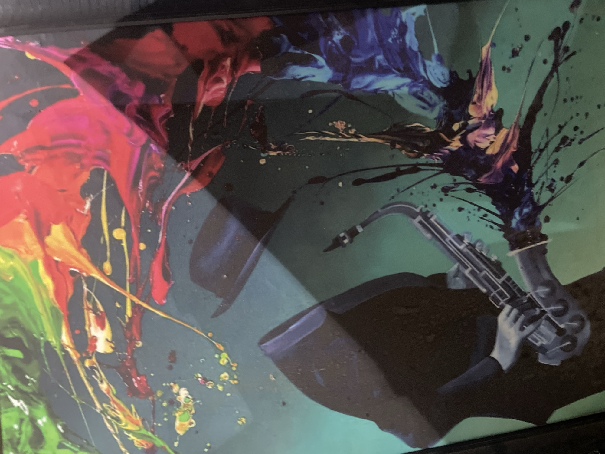 The image is a framed painting of a man playing a saxophone. The man is wearing a hat and suit, and the saxophone is depicted in grayscale. The background features splashes of vibrant colors including red, orange, yellow, green, and blue, creating an abstract, dynamic effect around the figure and instrument. The context indicates this piece of art is located somewhere in Abuja, Nigeria.