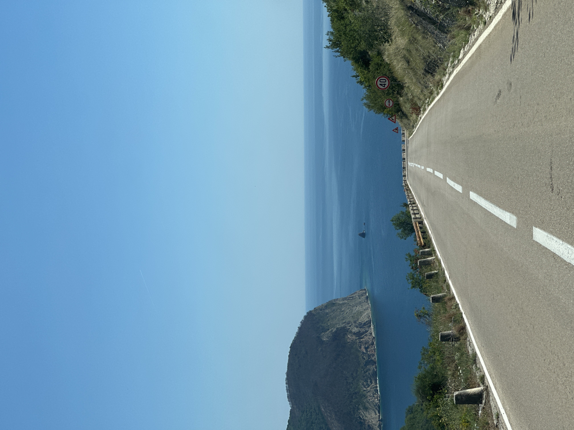 The image captures a coastal road near Novoselje, Montenegro, specifically a view of the Adriatic Sea. A paved road with white lane markings curves slightly downward. To the right of the road, the terrain slopes upwards with vegetation and road signs, including a speed limit sign with "40." To the left of the road, there's a drop-off with concrete posts and vegetation leading to the sea. An island or landmass protrudes into the blue sea. The sky is clear and blue. A faint jet stream contrail is visible in the upper left quadrant. A small boat is visible in the water.