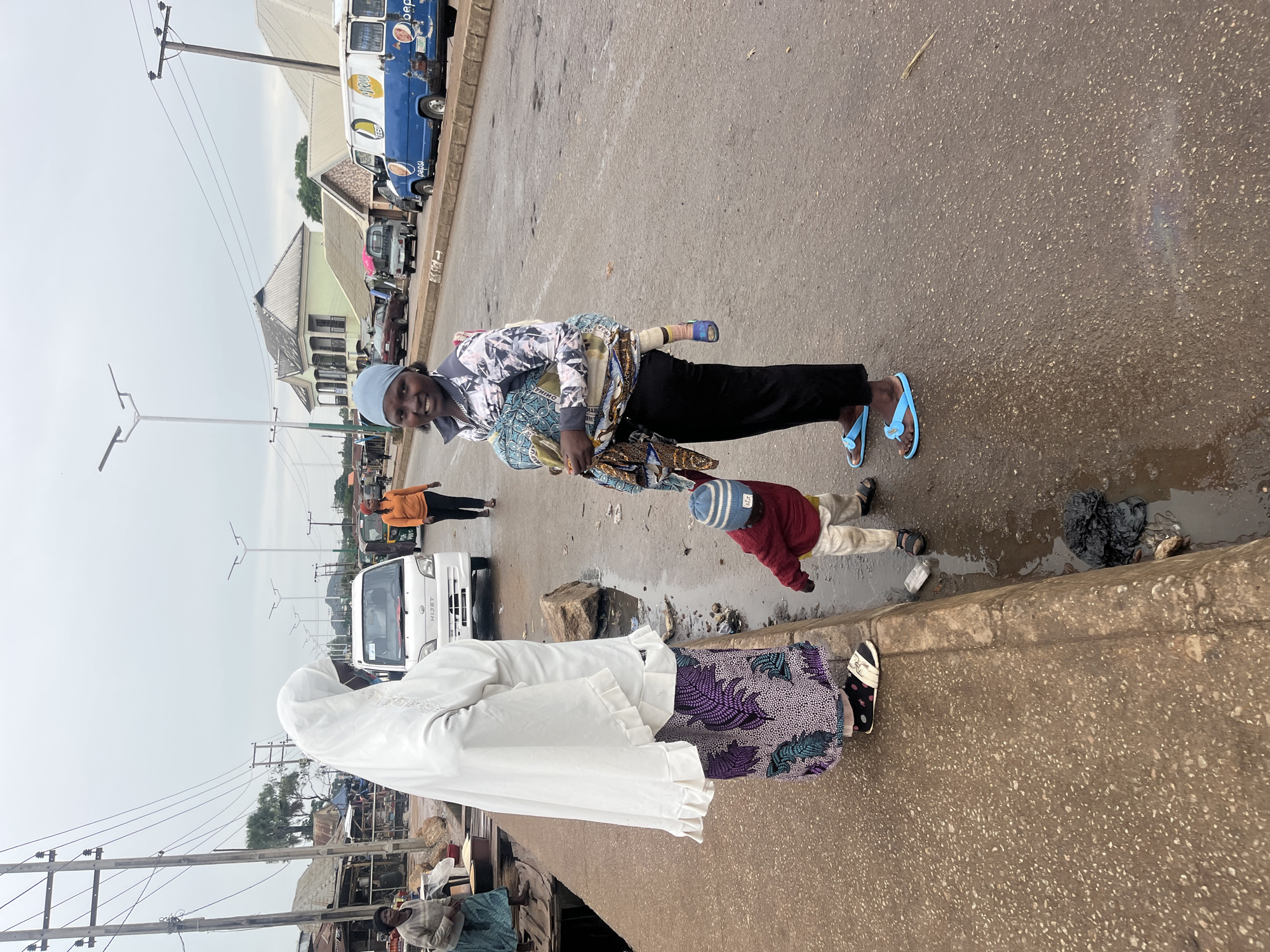 In Bukuru, Nigeria, on a grey, overcast day, a woman in a white hijab and a patterned skirt stands on a raised concrete walkway, facing a street. Next to her, a young child in a red sweater and white pants is looking down. To her right, another woman, wearing a light blue headscarf and a patterned shirt with black pants, is smiling and carrying a baby wrapped in a patterned cloth in a sling on her back. The street is paved and appears wet, with puddles and some trash, including a black plastic bag, visible near the curb. Several vehicles are present on the street, including a white Hiace van, a blue Pepsi van, and other cars. In the background, buildings with pitched roofs and overhead power lines are visible, suggesting an urban or semi-urban environment. A third woman in an orange shirt and dark pants is walking away from the camera on the street.
